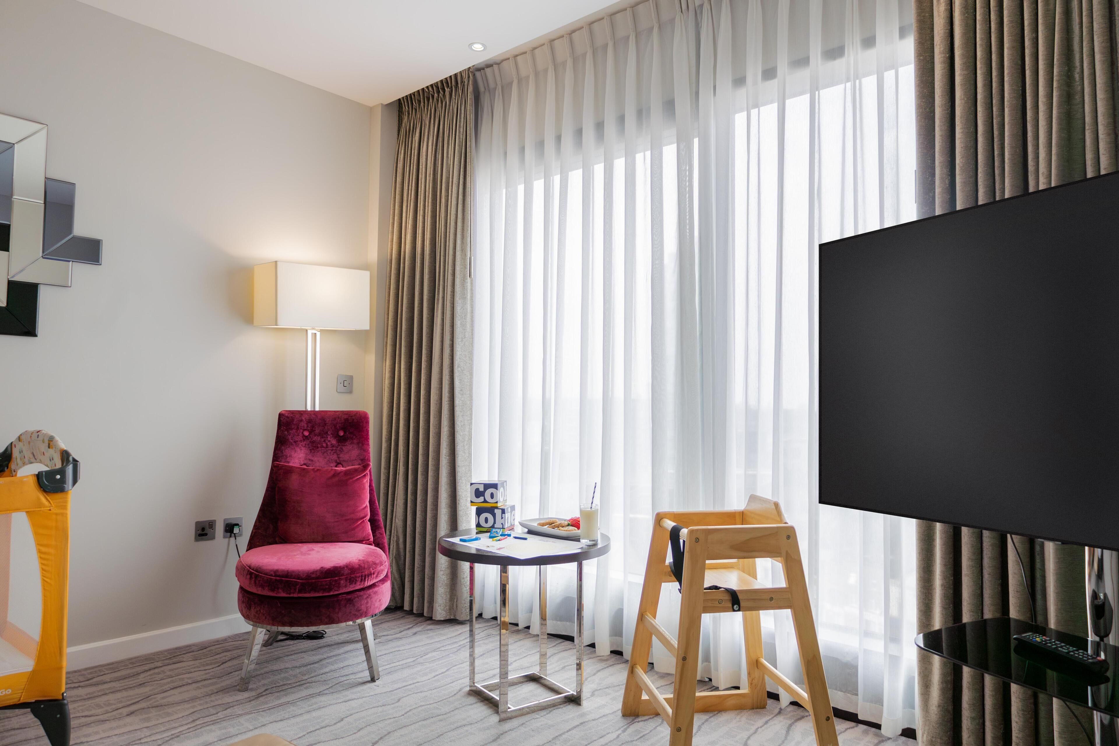 Una camera d'albergo con una grande TV a schermo piatto, una poltrona in velluto rosso, un piccolo tavolino con una lampada e una sedia di legno.
