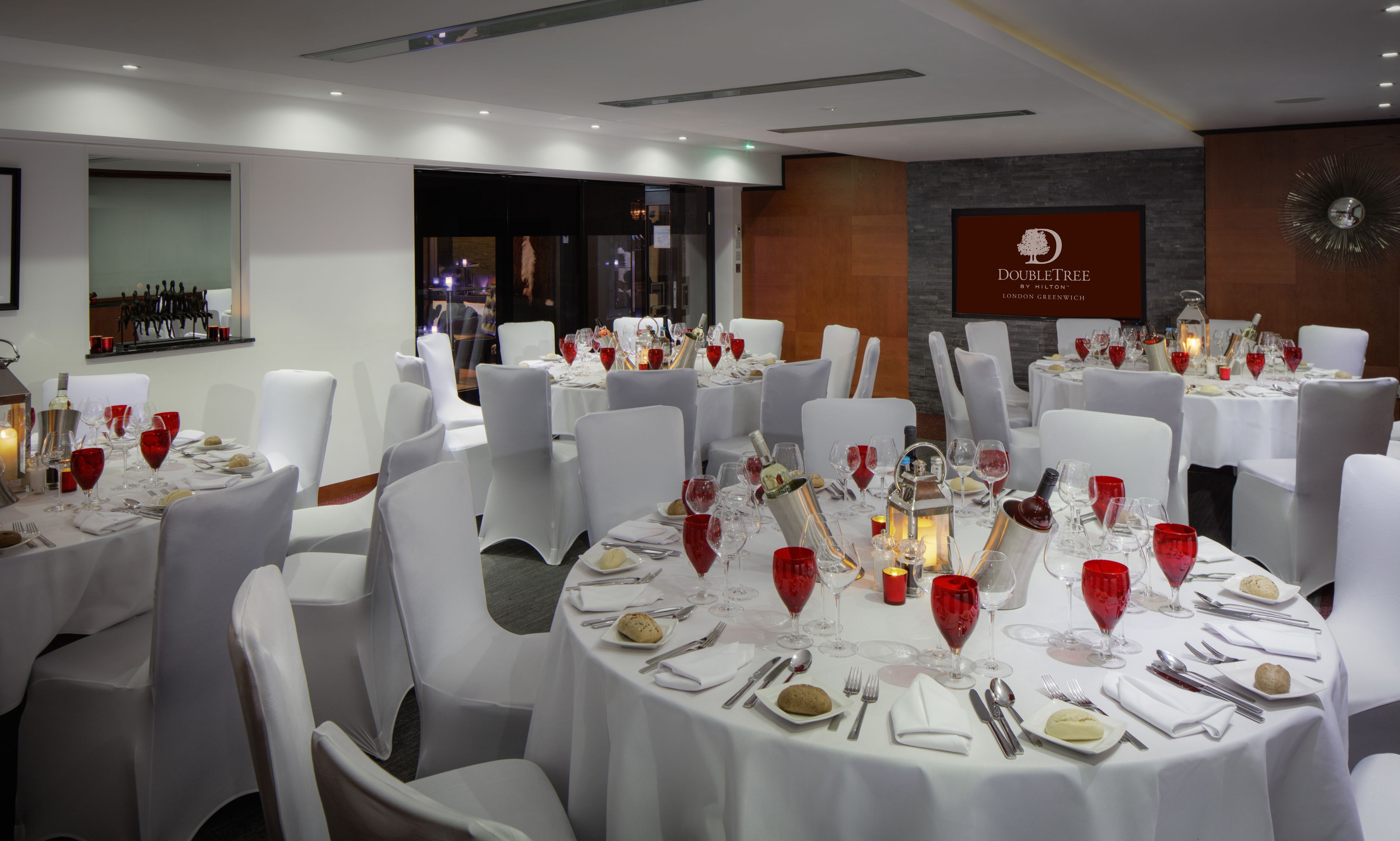 A well-lit, elegantly set dining area with round tables, white chairs, and red wine glasses.