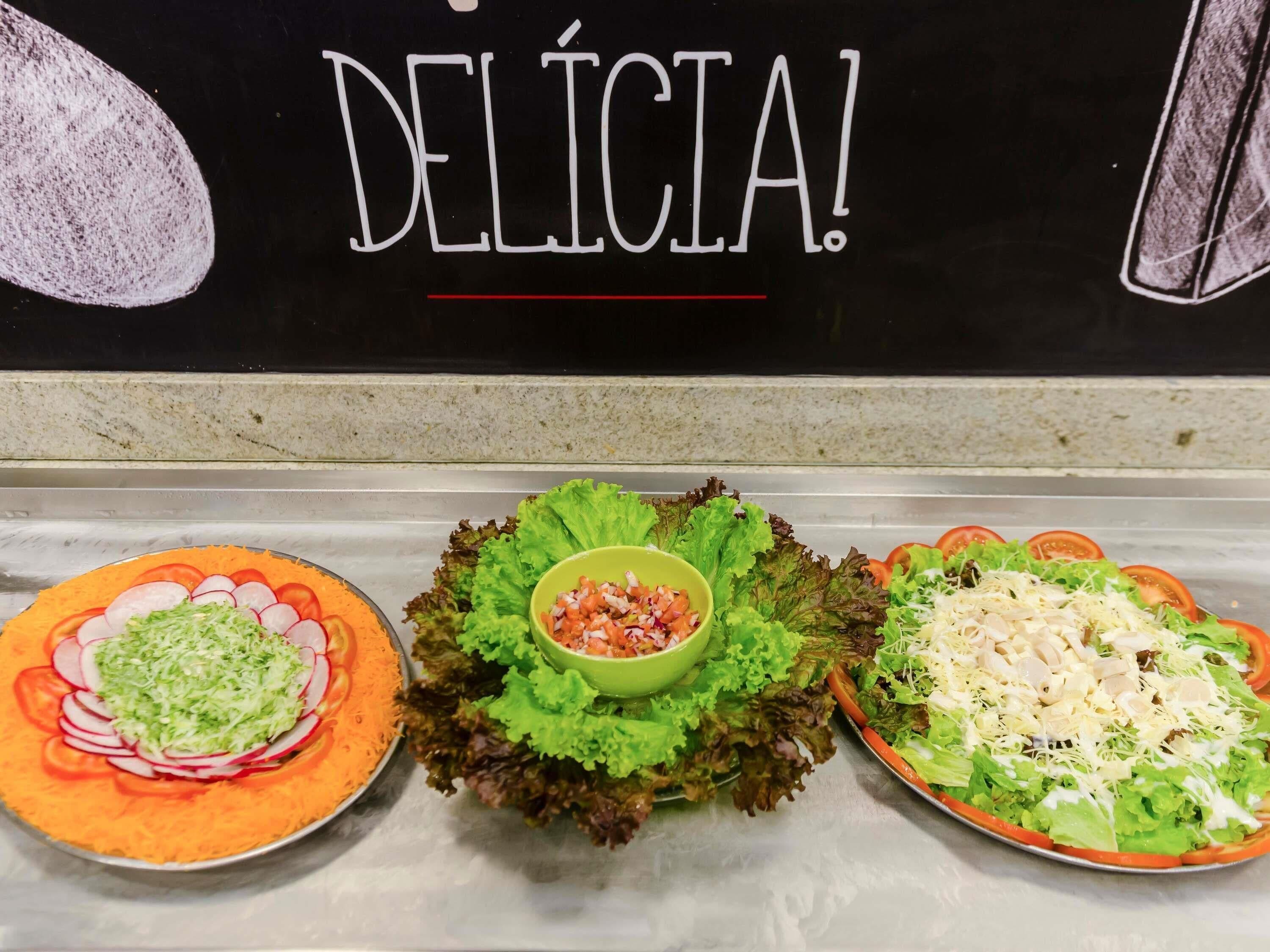 Tres platos de comida con lechuga, tomates y queso rallado, expuestos en una barra con un cartel que dice "DELICIA!"