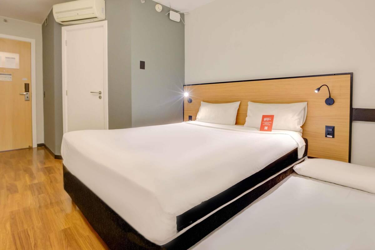 Habitación de hotel con una cama, cabecera de madera, mesita de noche, un reloj de pared y un equipo de aire acondicionado.