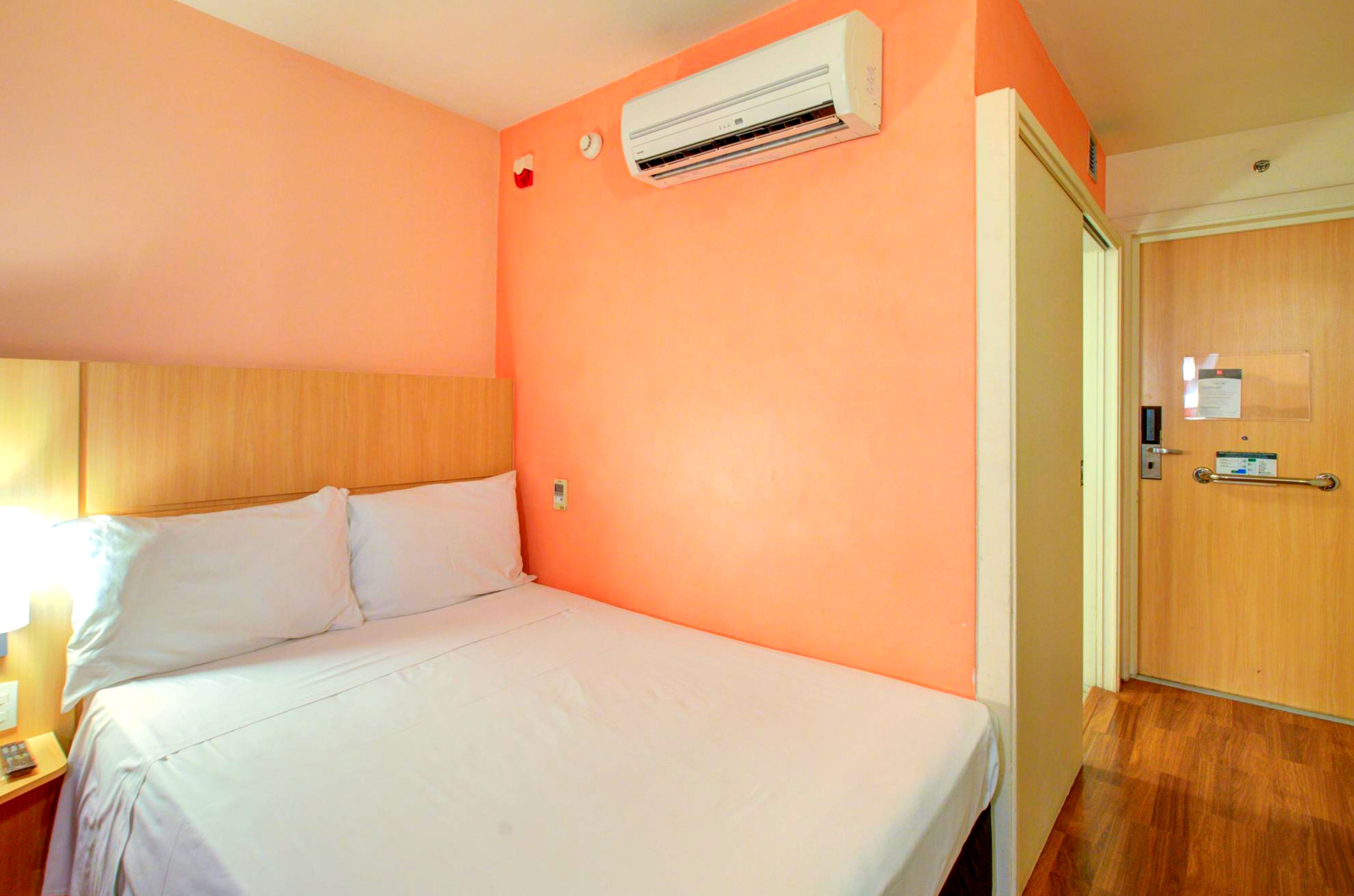 Una habitación de hotel con cama, aire acondicionado, una puerta de madera y una mesita de noche con lámpara.
