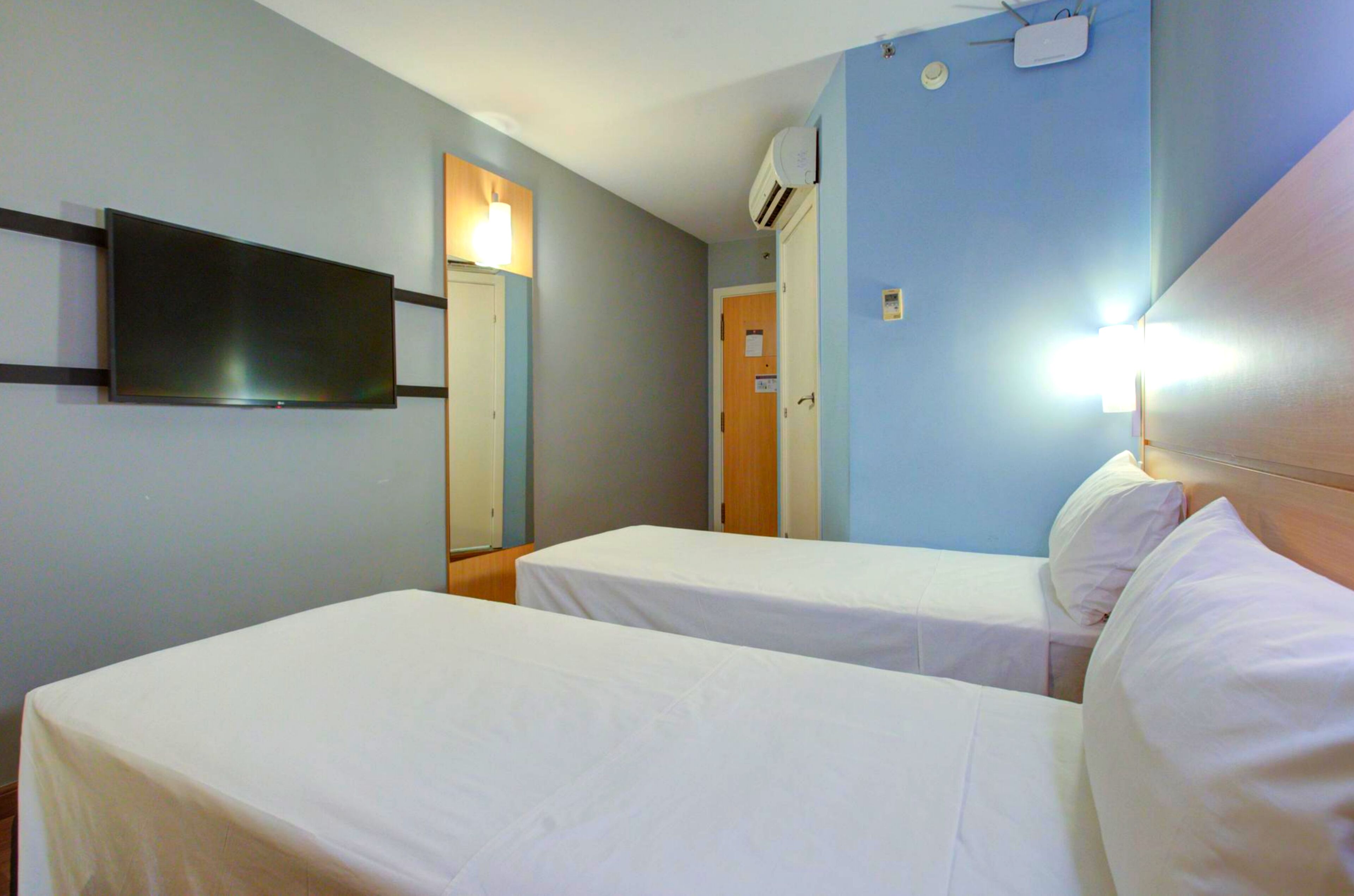 A hotel room with two single beds, a flat-screen TV mounted on the wall, and a bedside lamp.