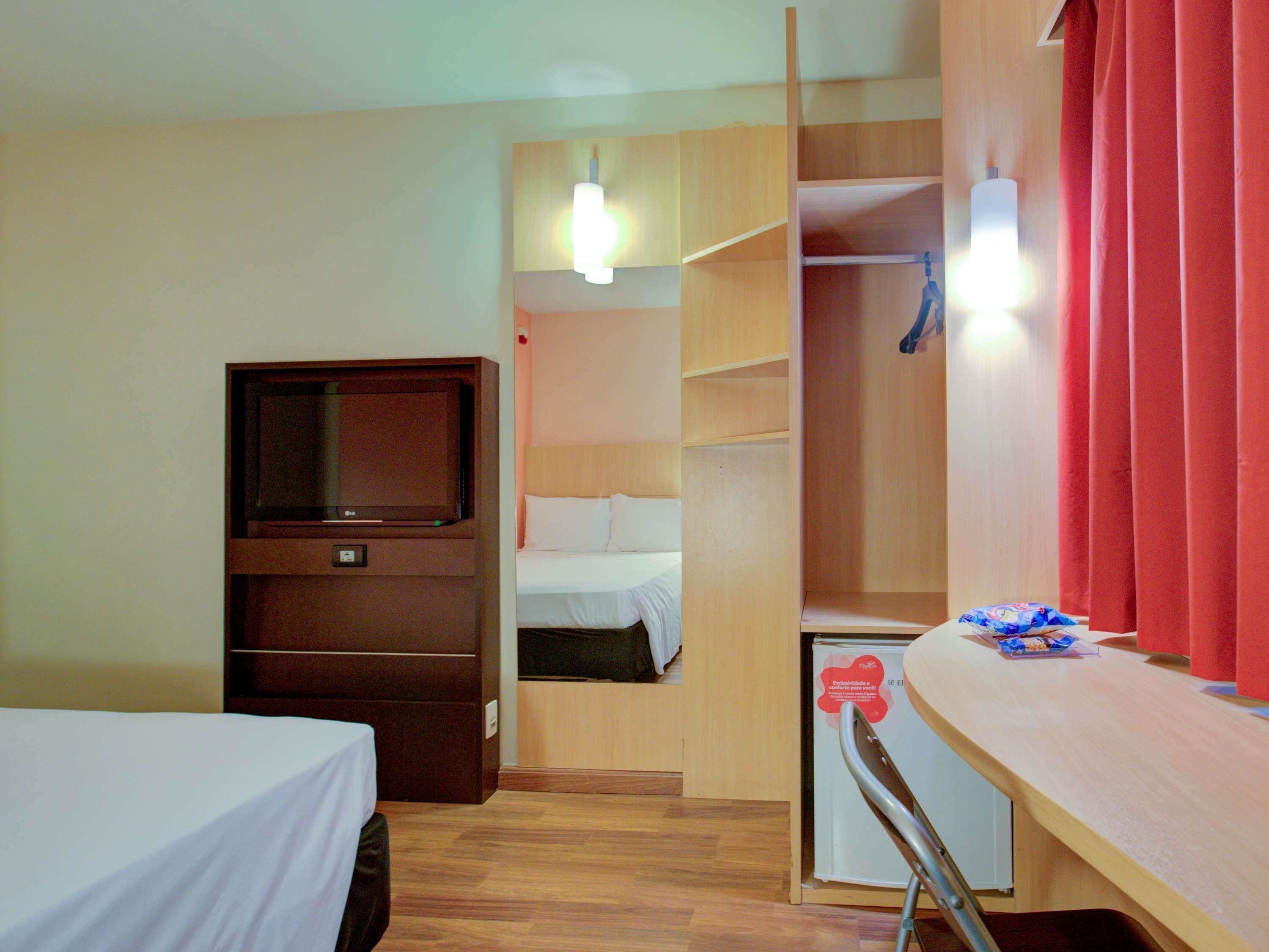 Habitación de hotel con cama, escritorio, televisión, armario y refrigerador pequeño.
