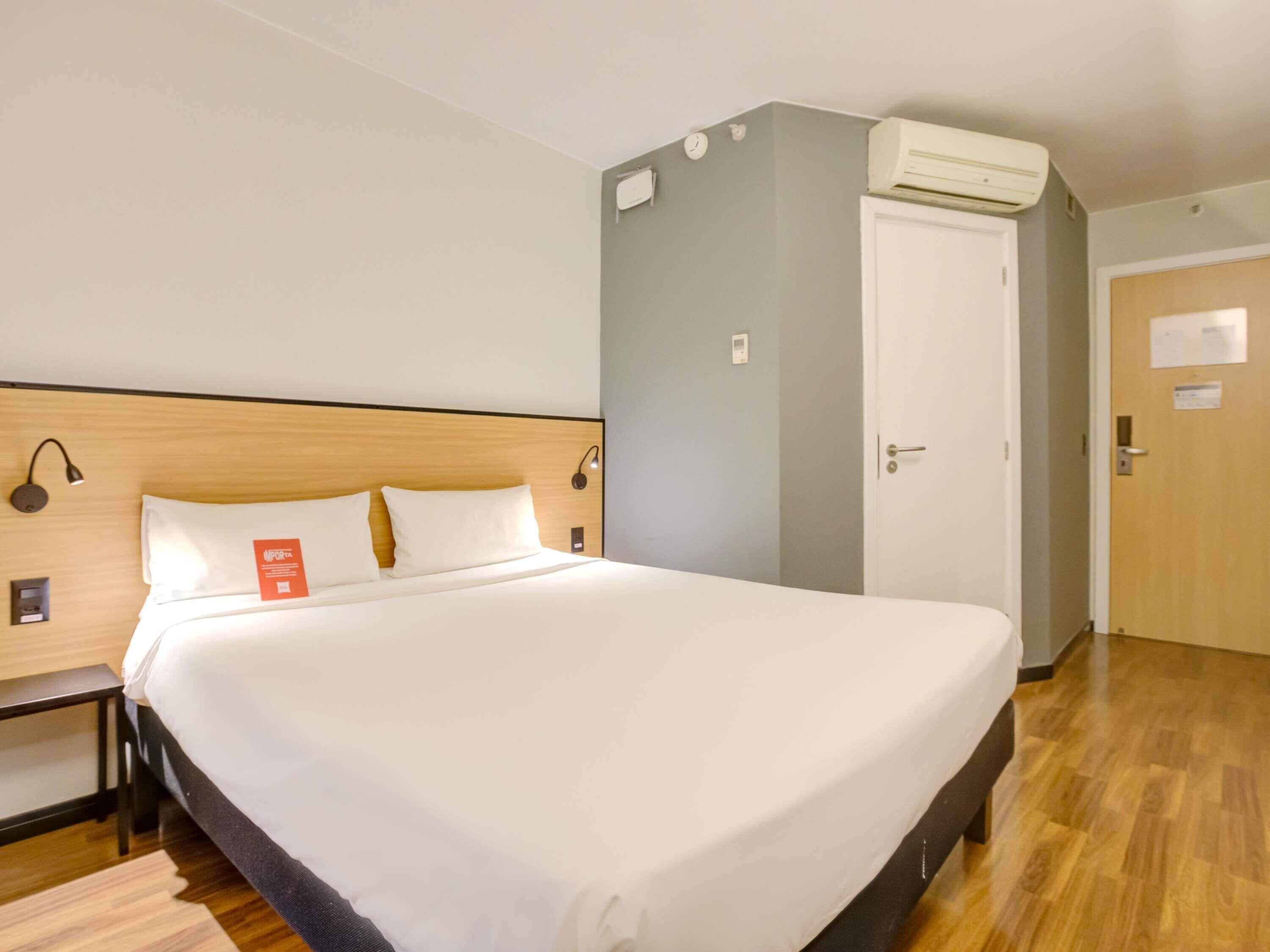 Habitación de hotel con una cama grande, cabecera de madera, mesita de noche, unidad de aire acondicionado, una puerta y un aplique de pared.