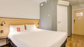 Habitación de hotel con una cama grande, cabecera de madera, mesita de noche, unidad de aire acondicionado, una puerta y un aplique de pared.