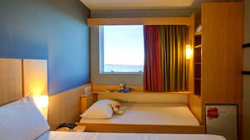 A hotel room with a bed, a bedside table, a lamp, a window with curtains, and a small refrigerator.