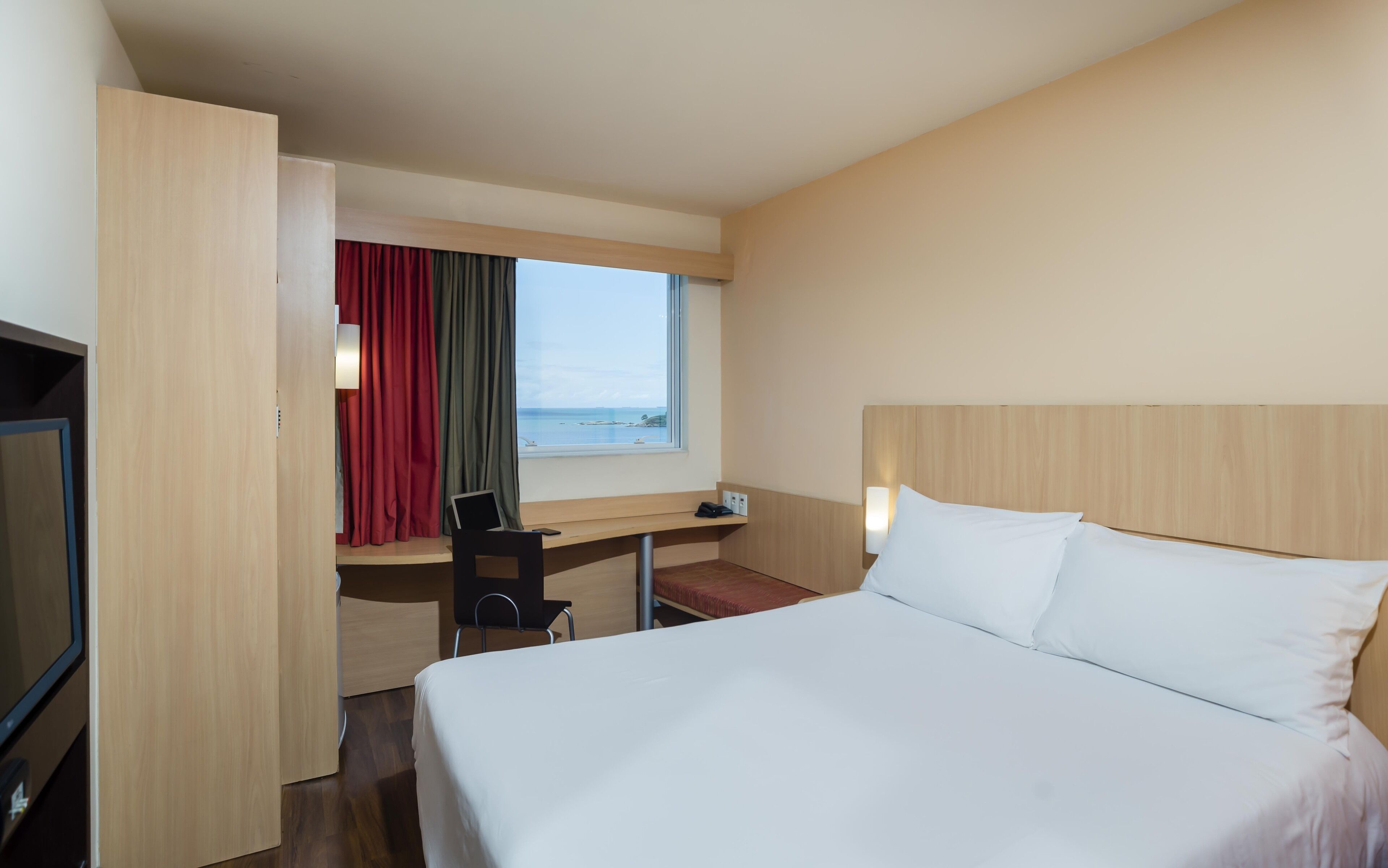A hotel room with a bed, a desk with a computer, a TV, and a window with curtains.