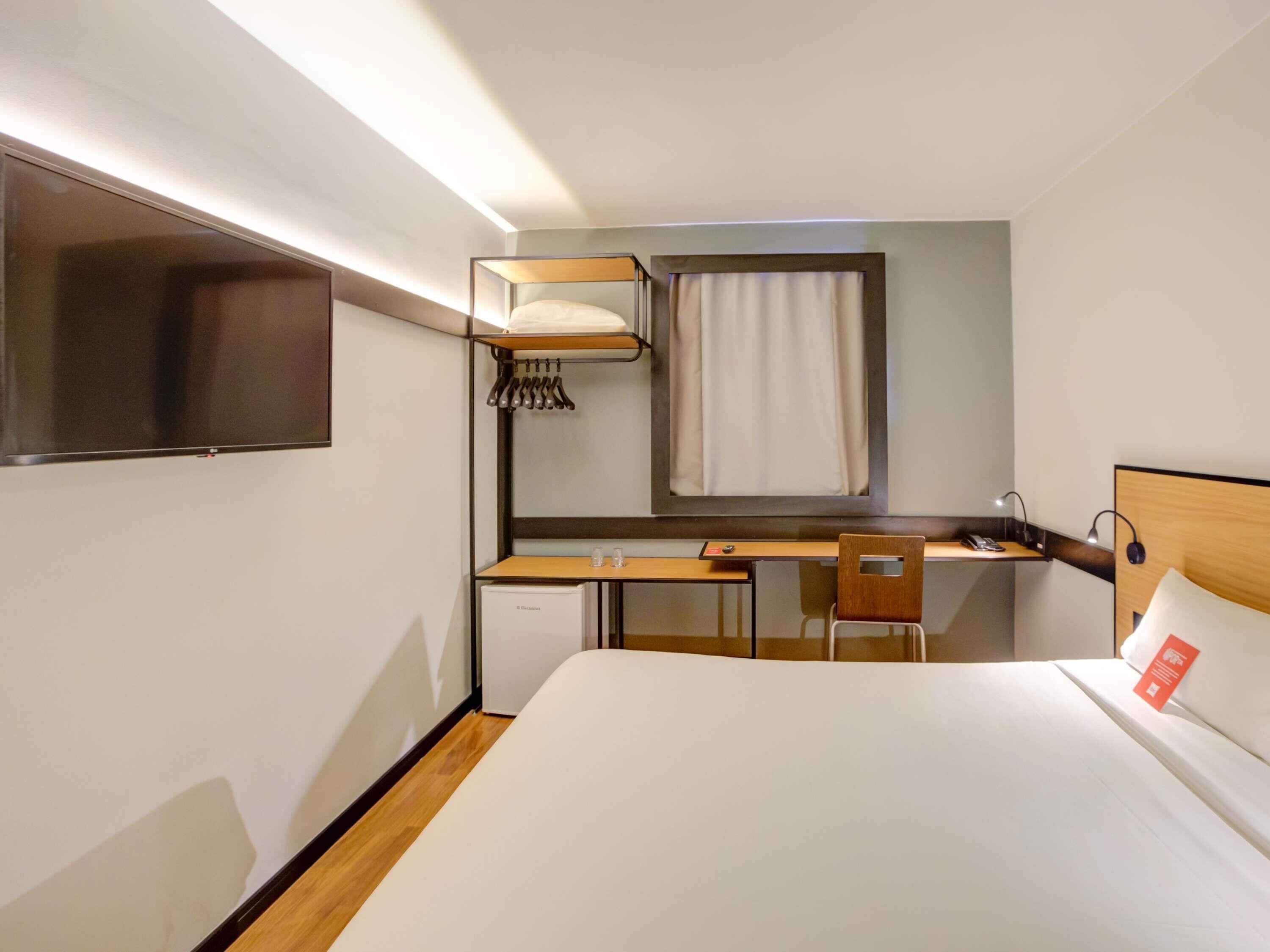 A hotel room with a bunk bed, a desk with a chair, a small refrigerator, a bedside table with a lamp, and a large mirror.