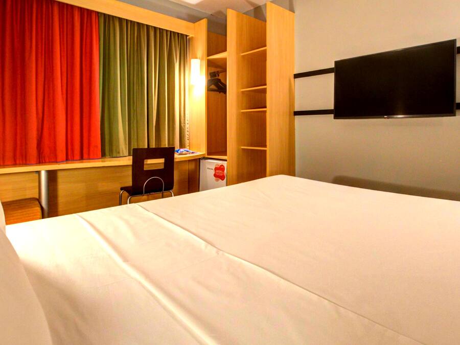 Ein Hotelzimmer mit Bett, Schreibtisch, Stuhl, an der Wand montiertem Fernseher und einer Garderobe mit Regalen.