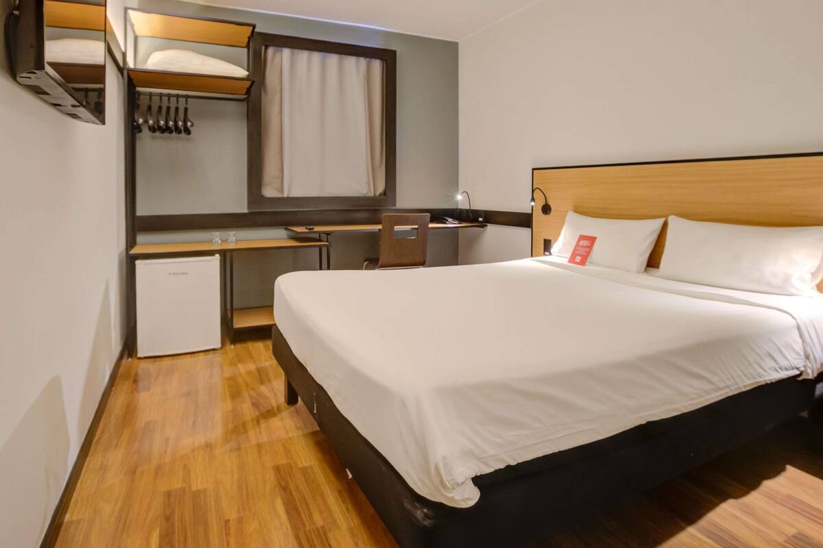 Una habitación de hotel con cama, escritorio, espejo y una pequeña kitchenette.
