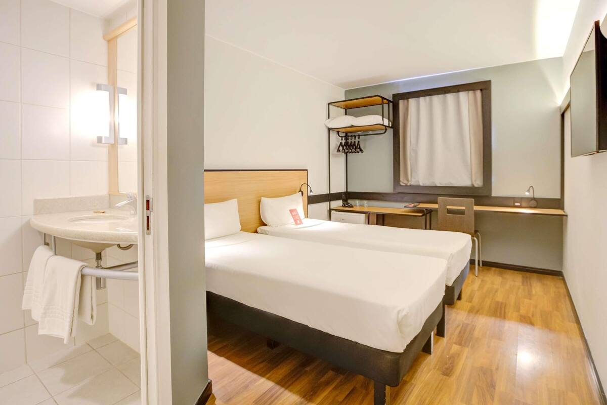 Habitación de hotel con cama doble, mesita de noche, escritorio, espejo, estante pequeño y baño con lavamanos y toallero.