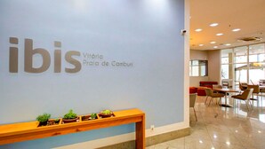 Un interior moderno con una pared que muestra 'ibis' y 'Vitória Praia de Camburi'. Hay un banco de madera con pequeñas plantas y una zona de estar con mesas y sillas.