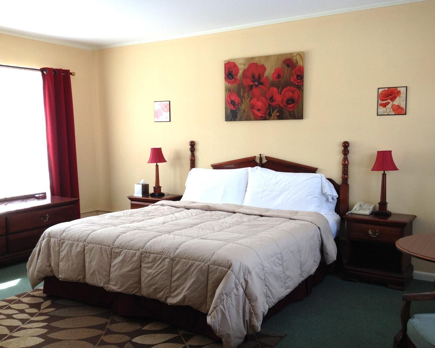 Une chambre d’hôtel avec un grand lit, deux tables de chevet avec des lampes, un tableau représentant des fleurs rouges accroché au mur et une fenêtre avec des rideaux rouges.