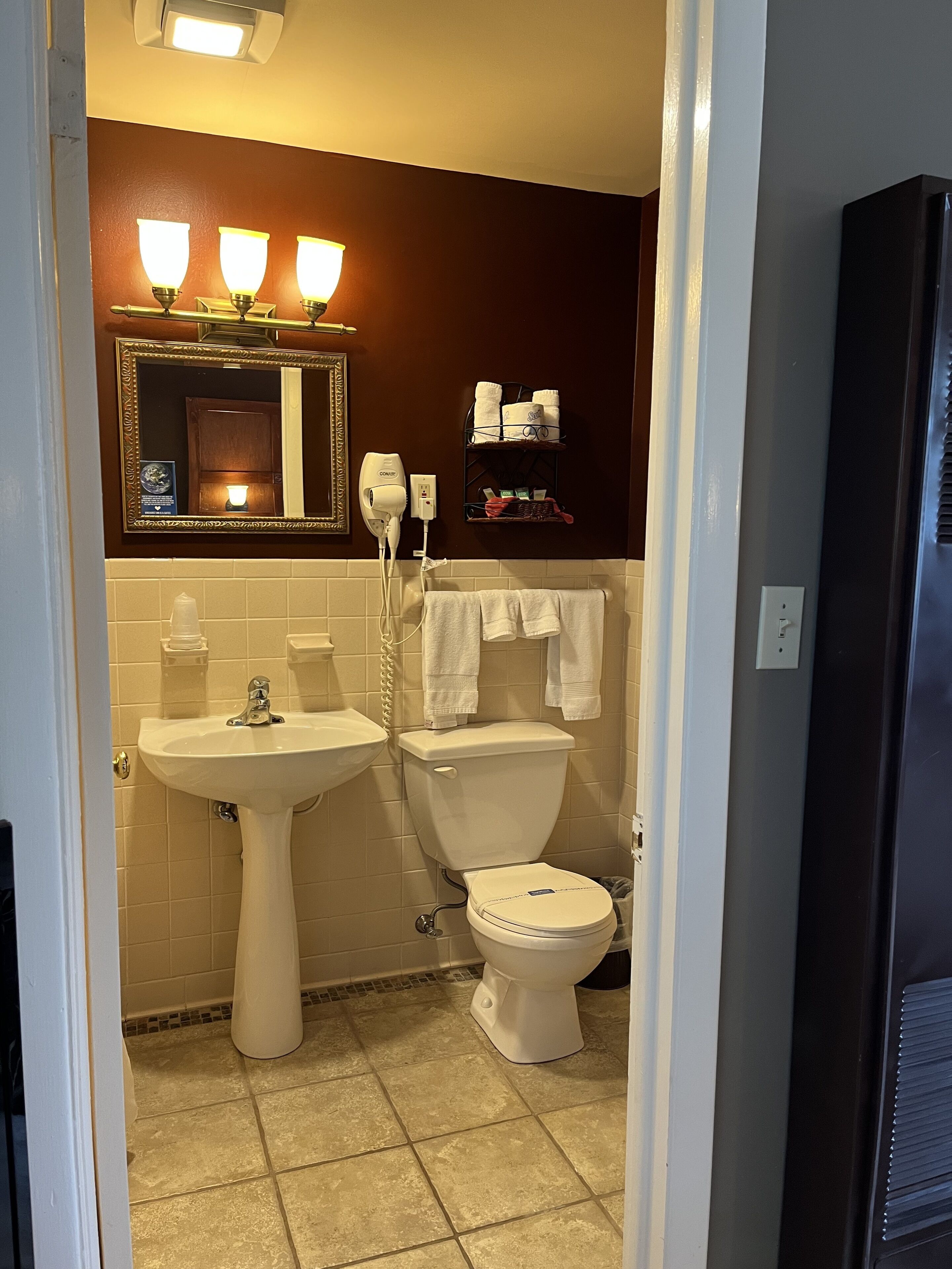 Ein Badezimmer mit einem Standwaschbecken, Toilette und einem Schminkspiegel. Über dem Spiegel sind drei Wandleuchten angebracht, und es gibt einen Handtuchhalter mit Handtüchern.