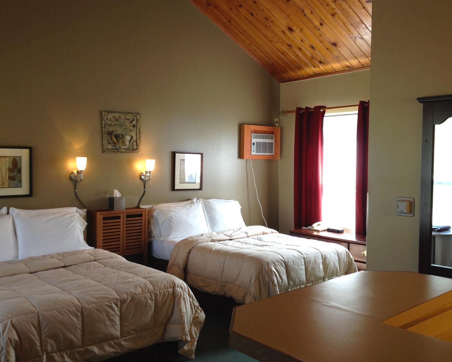 A hotel room with two beds, a wooden ceiling, a window with red curtains, and wall-mounted lights.
