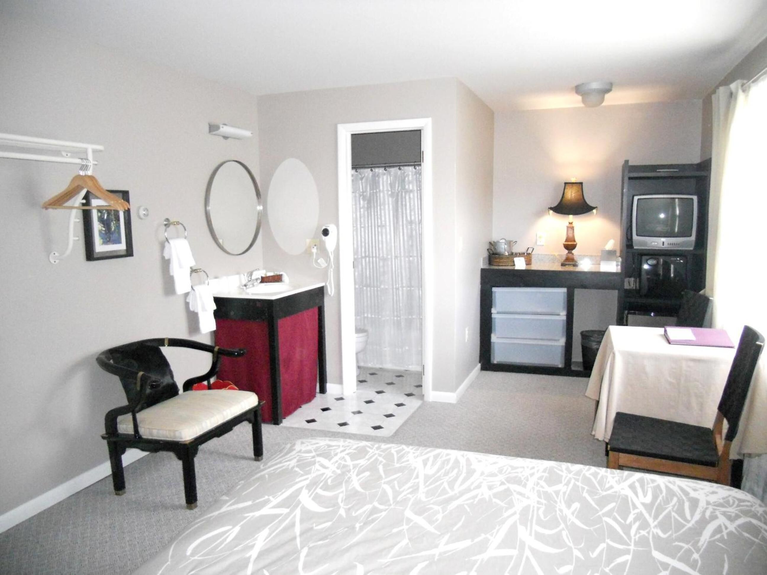 A compact hotel room with a bed, a desk, a chair, a small kitchenette, and a TV.