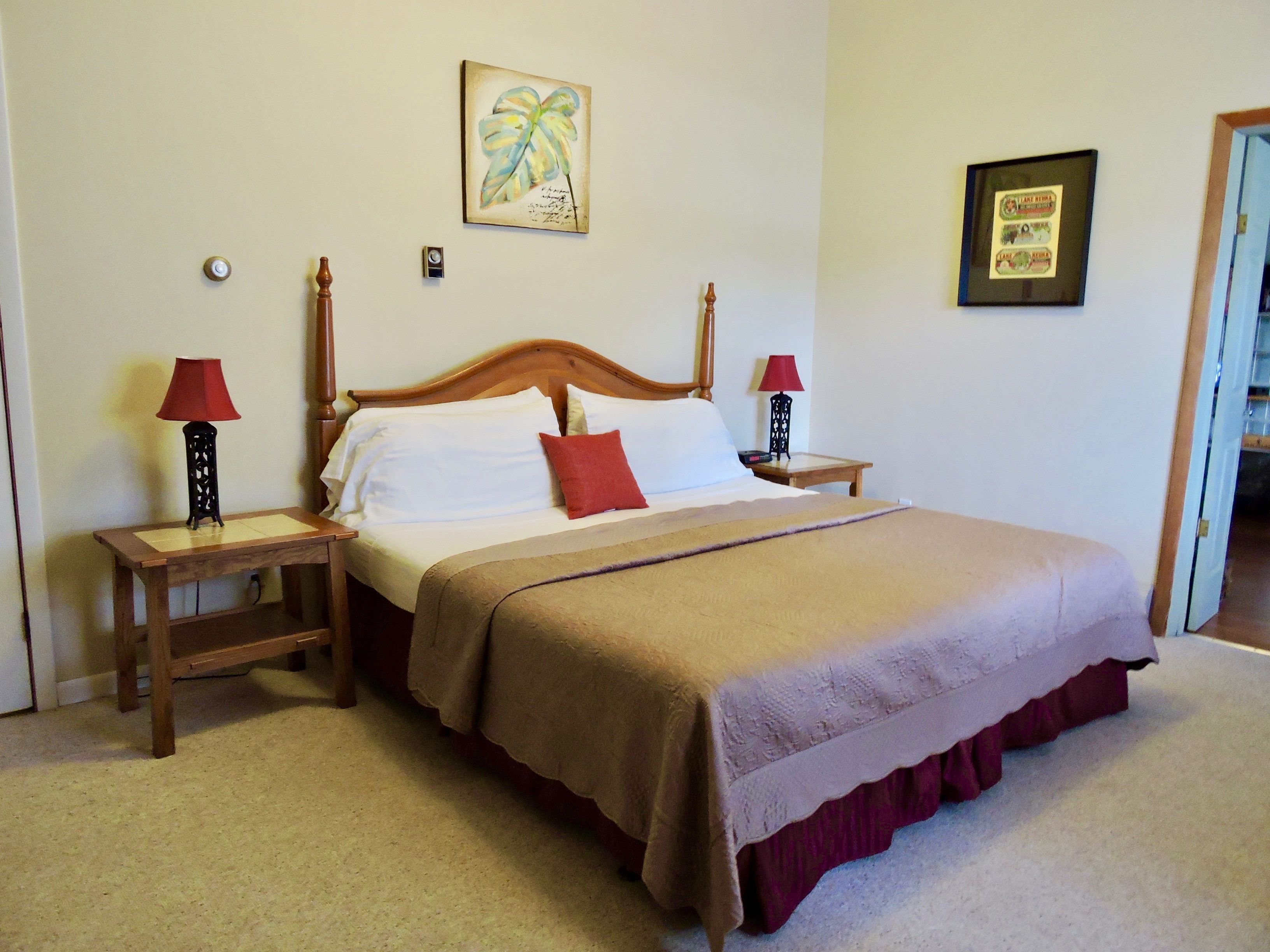 Cama bem arrumada com cabeceira de madeira, duas mesas de cabeceira com abajures, um travesseiro decorativo vermelho e um quadro na parede.