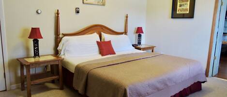 Cama bem arrumada com cabeceira de madeira, duas mesas de cabeceira com abajures, um travesseiro decorativo vermelho e um quadro na parede.