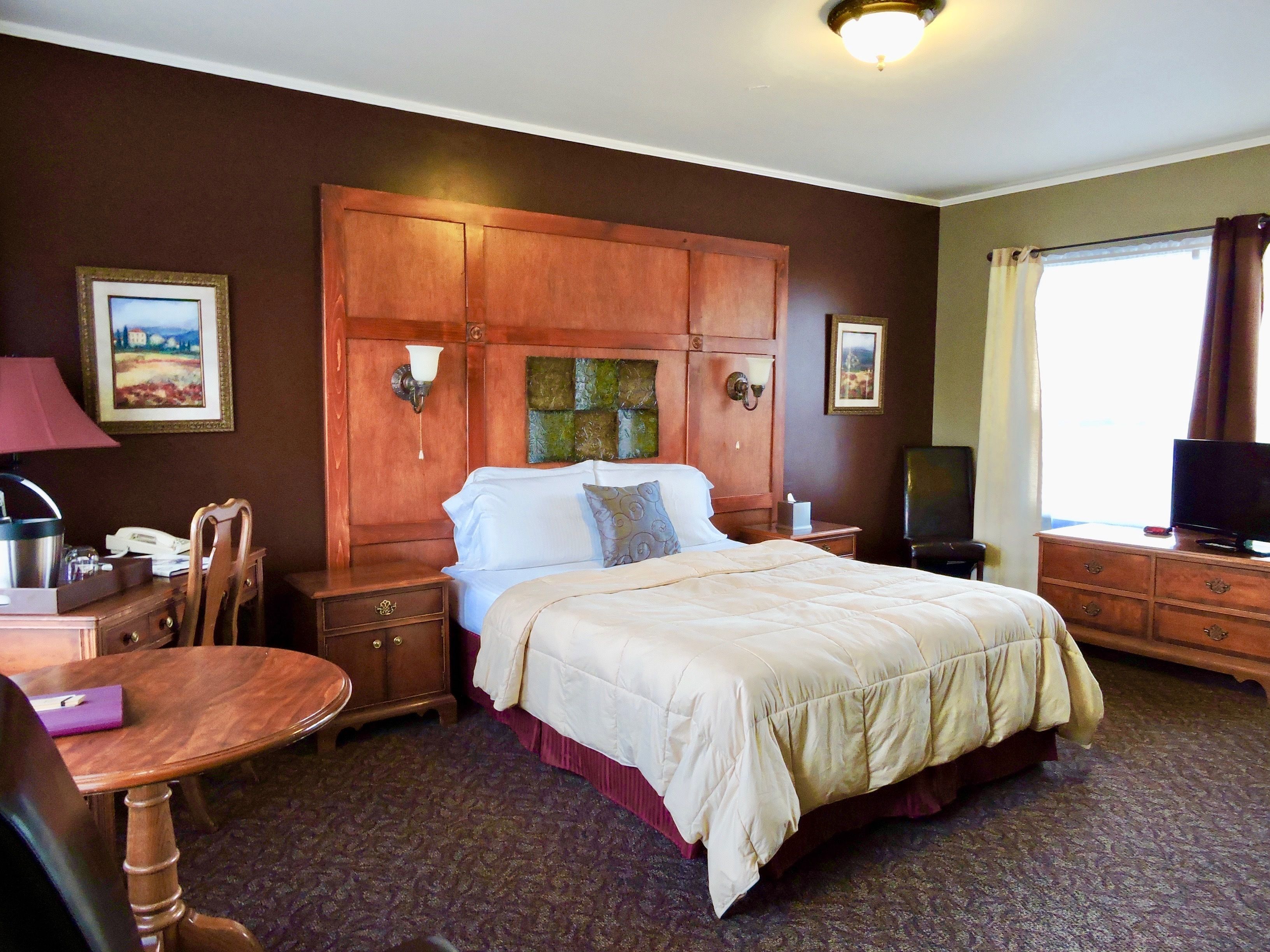 A hotel room with a large bed, wooden headboard, a desk with a chair, a television, and a window with curtains.