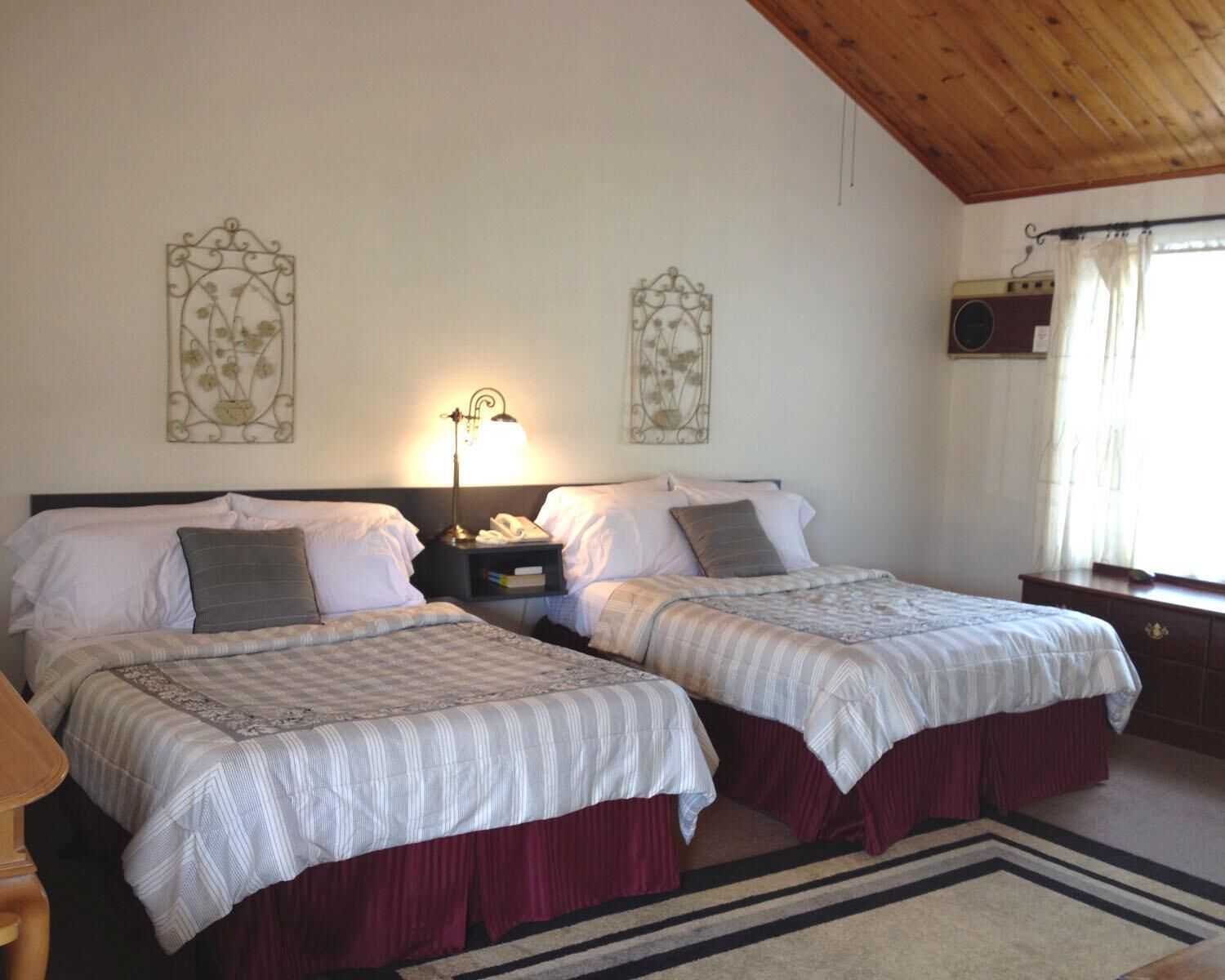 Habitación doble con dos camas, cada una con una colcha a rayas y una falda roja. Entre las camas hay una mesita de noche con una lámpara, y una pequeña mesa con un teléfono encima. La habitación tiene techo de madera y una ventana con cortinas.