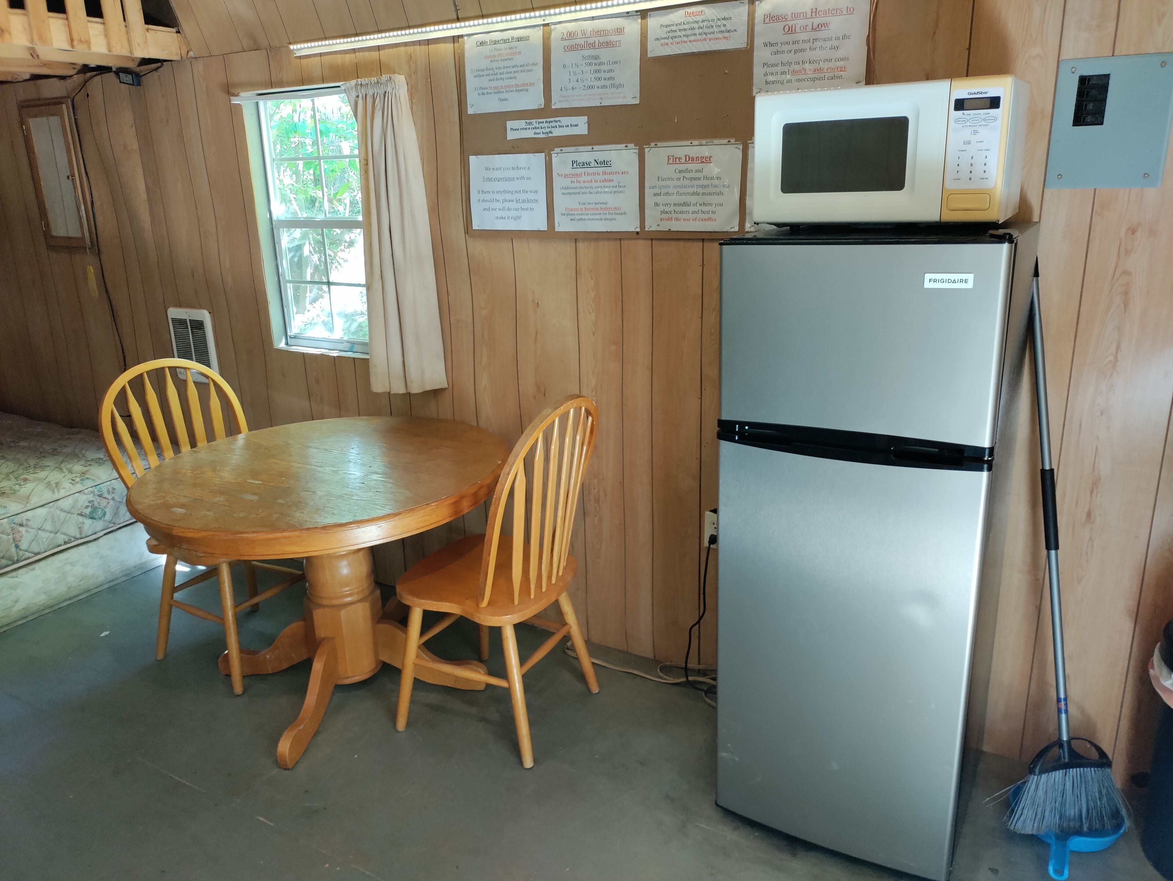 A wooden cabin interior with a dining table, two chairs, a refrigerator, a microwave, and various informational signs on the walls.