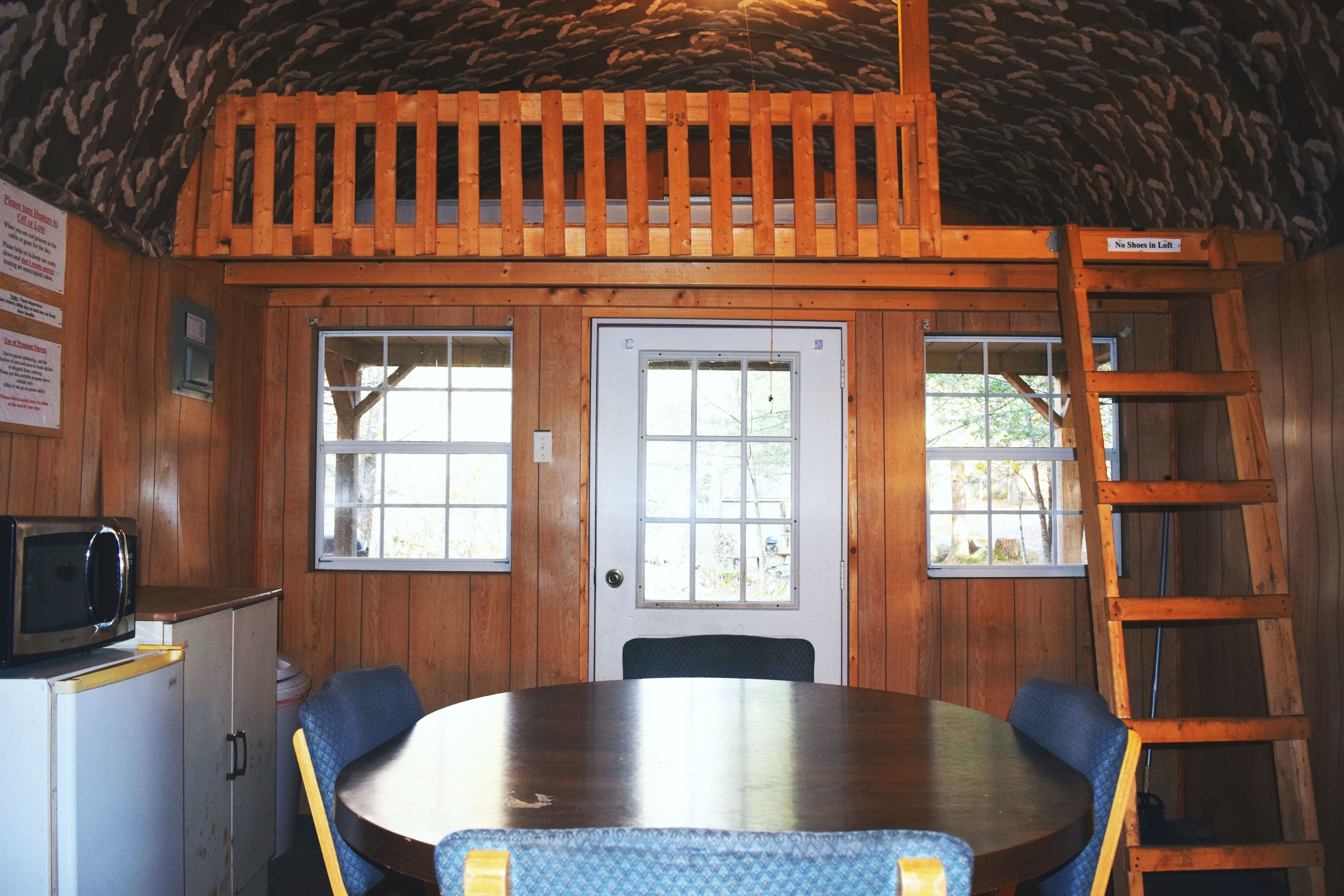 A wooden cabin with a dining table, two chairs, a microwave, and a ladder.