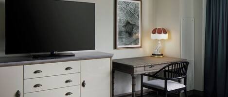 A hotel room with a flat-screen TV, a desk with a chair, a framed map on the wall, and a lamp.