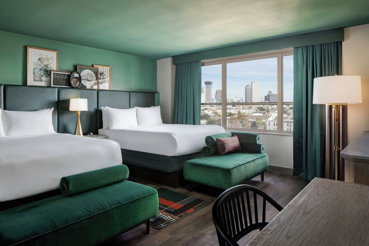A hotel room with two beds, a green armchair, a dining table, and a view of the cityscape.