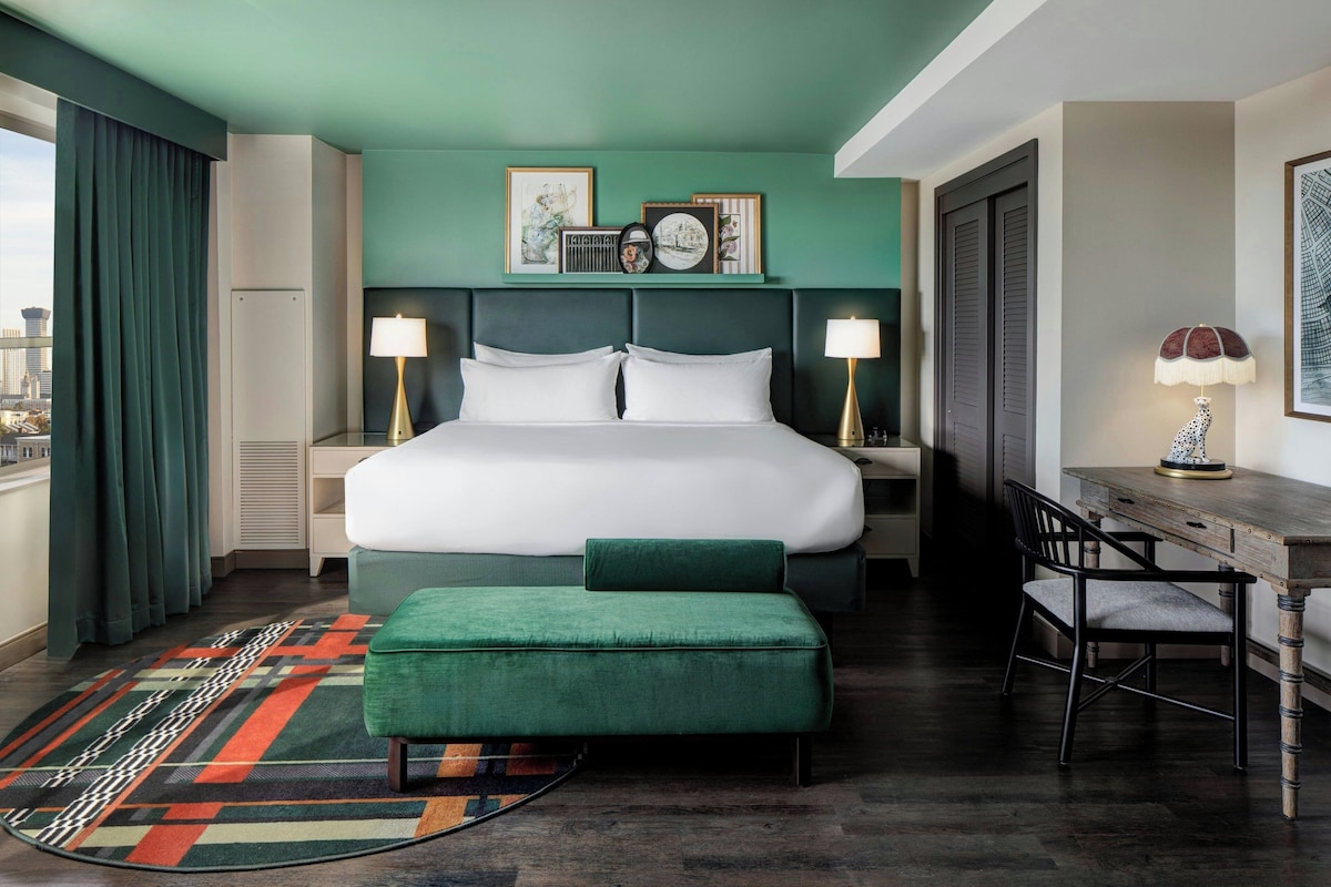 Une chambre d’hôtel avec un grand lit, deux tables de chevet avec des lampes, un bureau avec une chaise, un pouf vert, un tapis coloré et une vue sur la ville par la fenêtre.