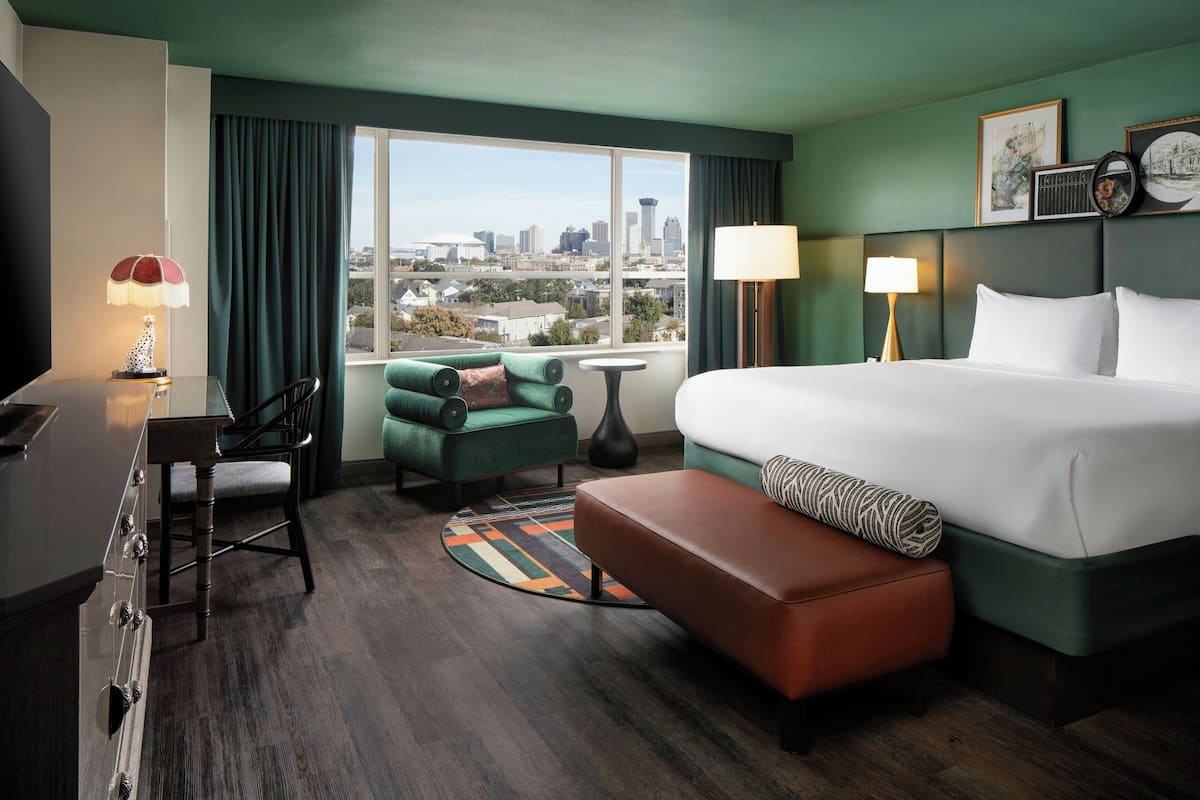 A hotel room with a large bed, a desk, a chair, a small table, and a view of the city skyline.