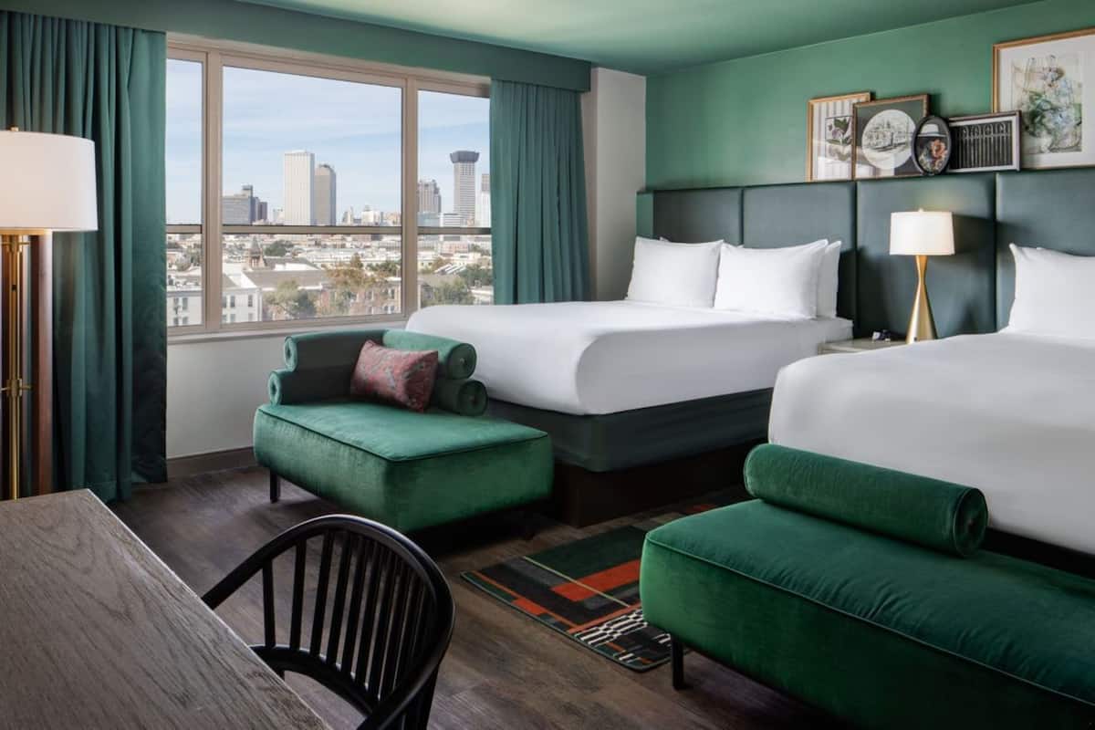 A hotel room with two beds, a desk, and a chair. There is a view of a cityscape through the window.