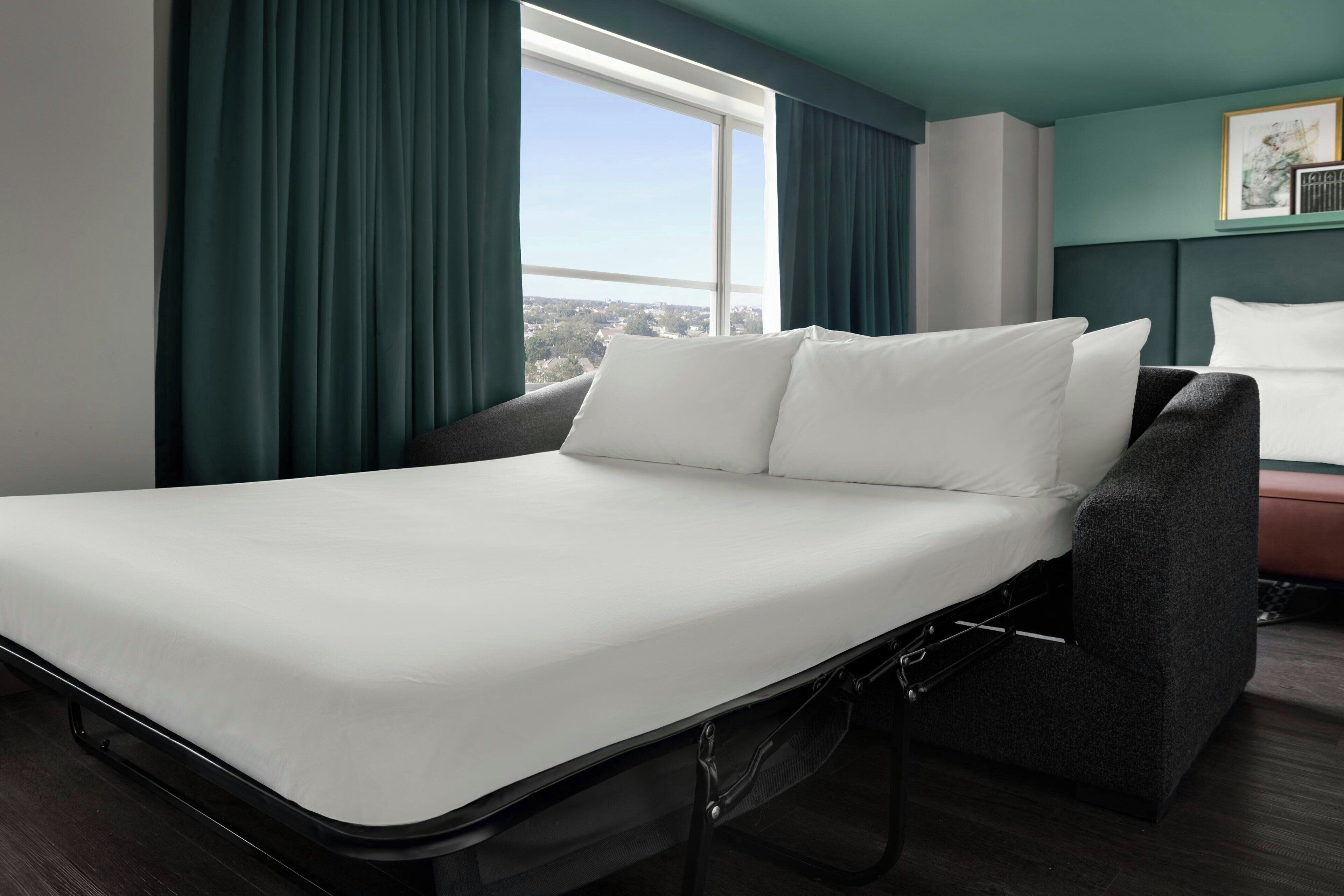 A modern hotel room with a bed, a chair, a window with curtains, and a view of the city.