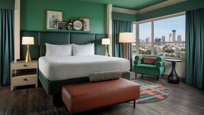 Een hotelkamer met een groot bed, twee nachtkastjes, een groene fauteuil, een ronde salontafel en uitzicht op de stadshemel.
