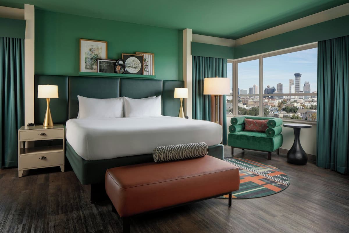 A hotel room with a large bed, two bedside tables, a green armchair, a round coffee table, and a view of the city skyline.