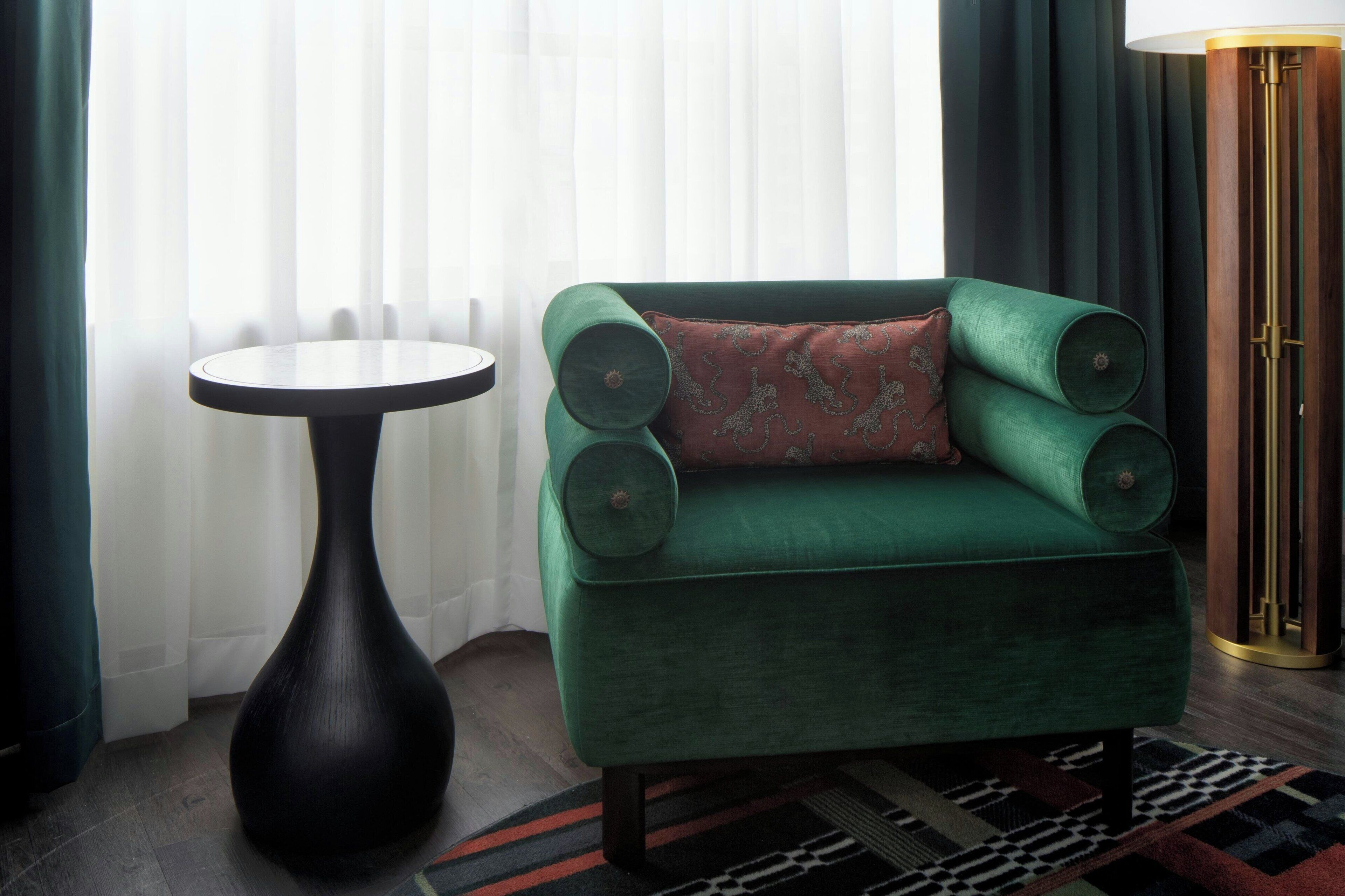 A green armchair with a patterned cushion, a round black side table, and a tall floor lamp with a gold base.