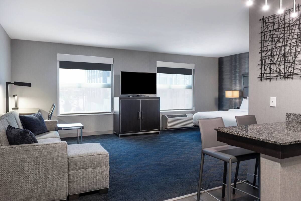 Ein modernes Hotelzimmer mit großem Flachbildfernseher, einer Couch, einem Essbereich und einem großen Fenster mit Blick auf die Stadt.