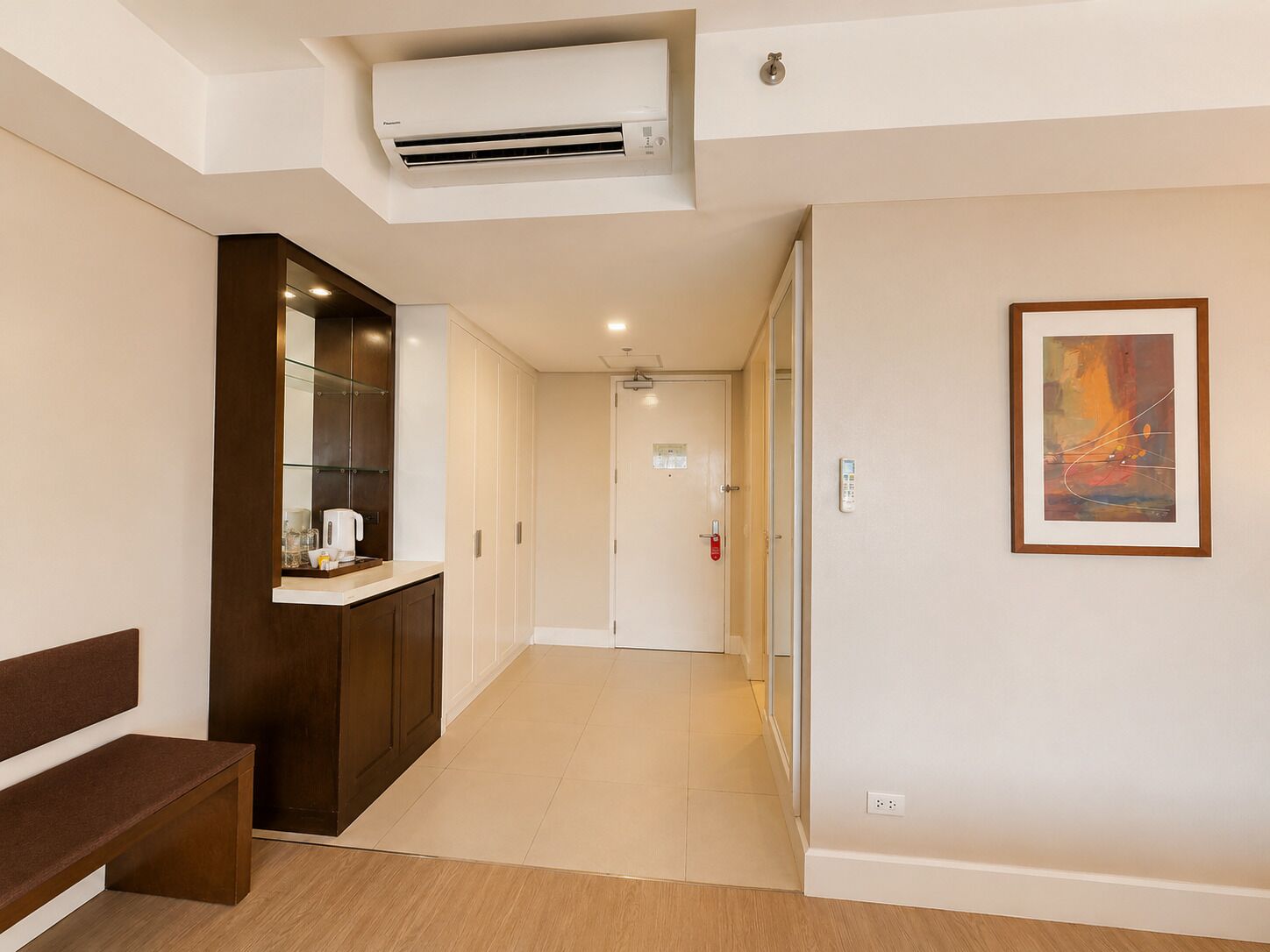 Un pasillo de hotel con un banco, una máquina de café, un equipo de aire acondicionado, un detector de incendios y un cuadro abstracto enmarcado.