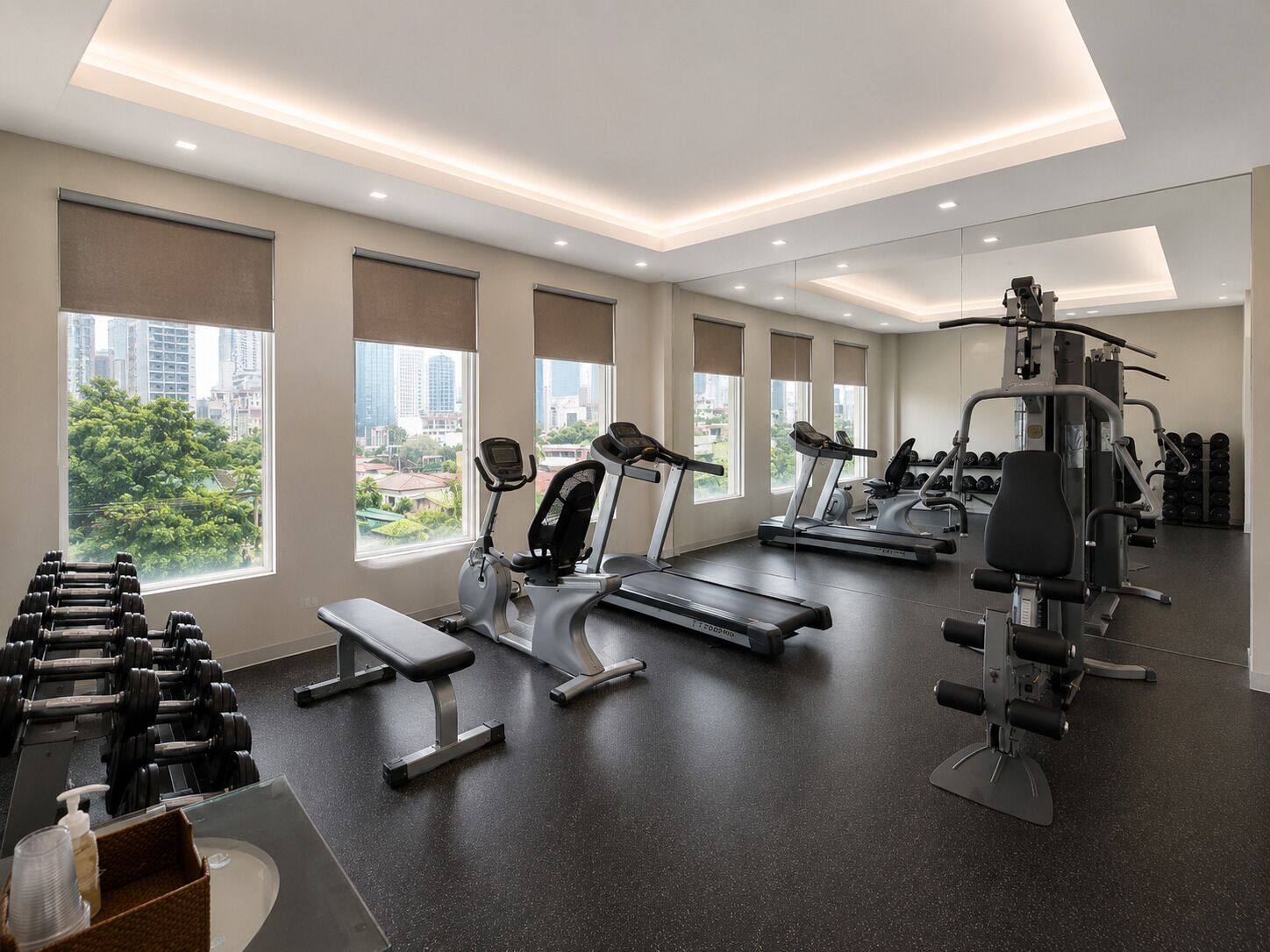 Un gimnasio moderno con cintas de correr, mancuernas y máquinas de pesas, amplias ventanas con vistas a la ciudad y un interior limpio y bien iluminado.