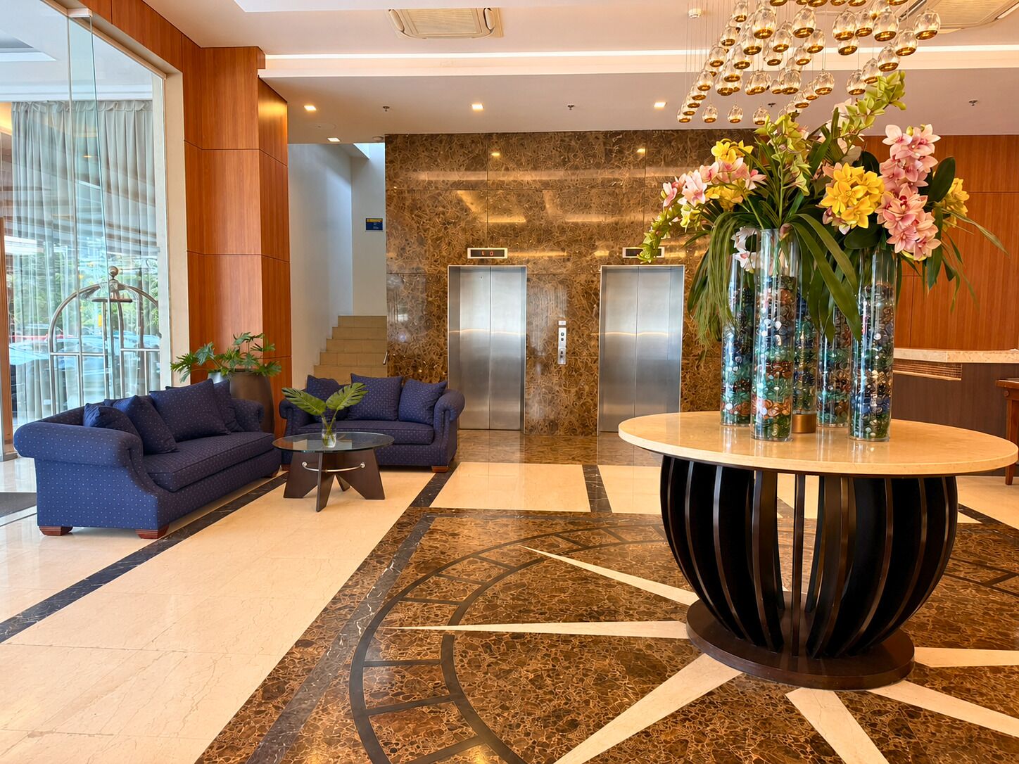 A lobby with a marble floor, two sofas, a round table with vases, and an elevator in the background.