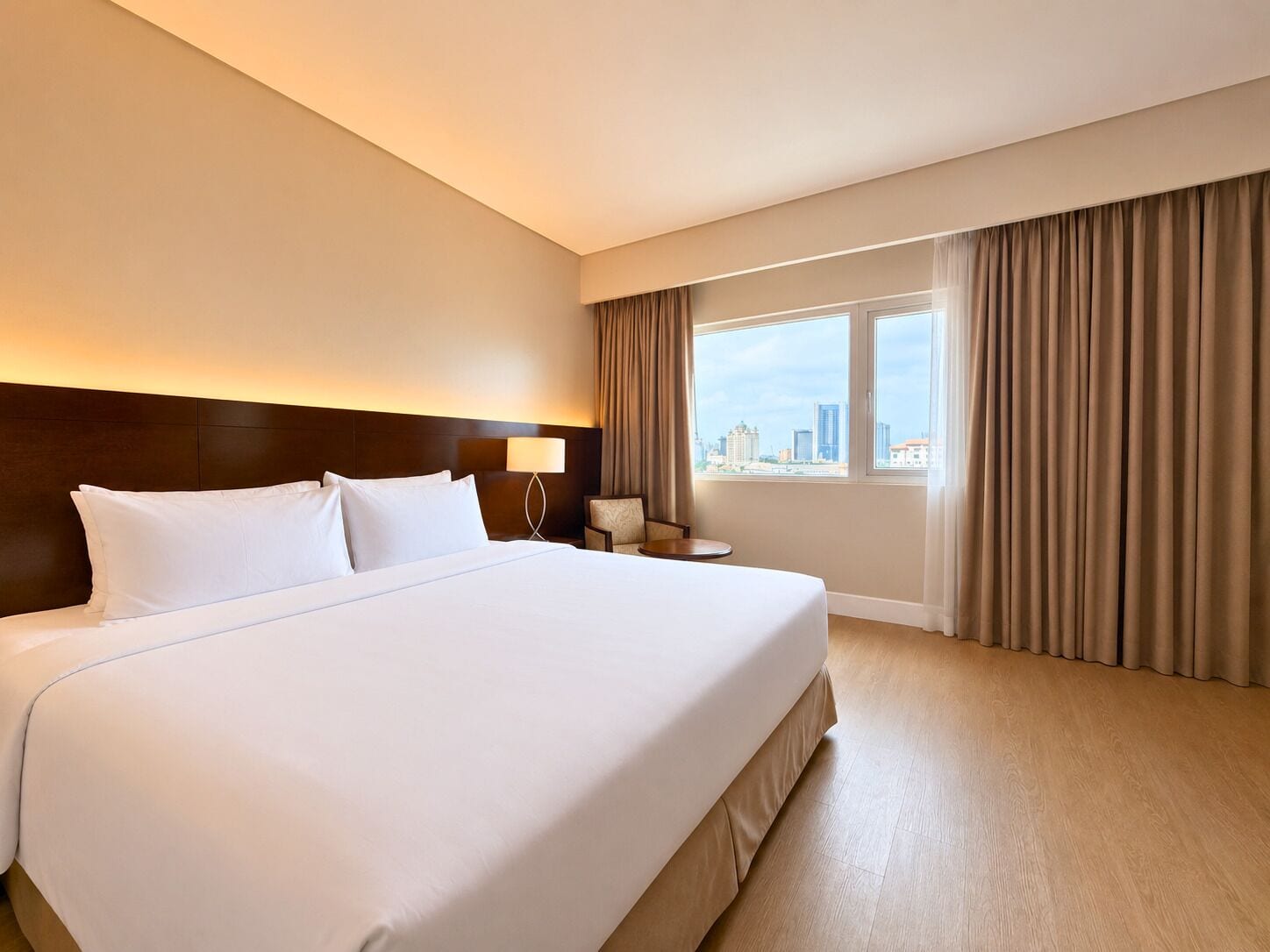Een hotelkamer met een groot bed, een nachtkastje, een lamp, een stoel, een raam met gordijnen en uitzicht op de stad.