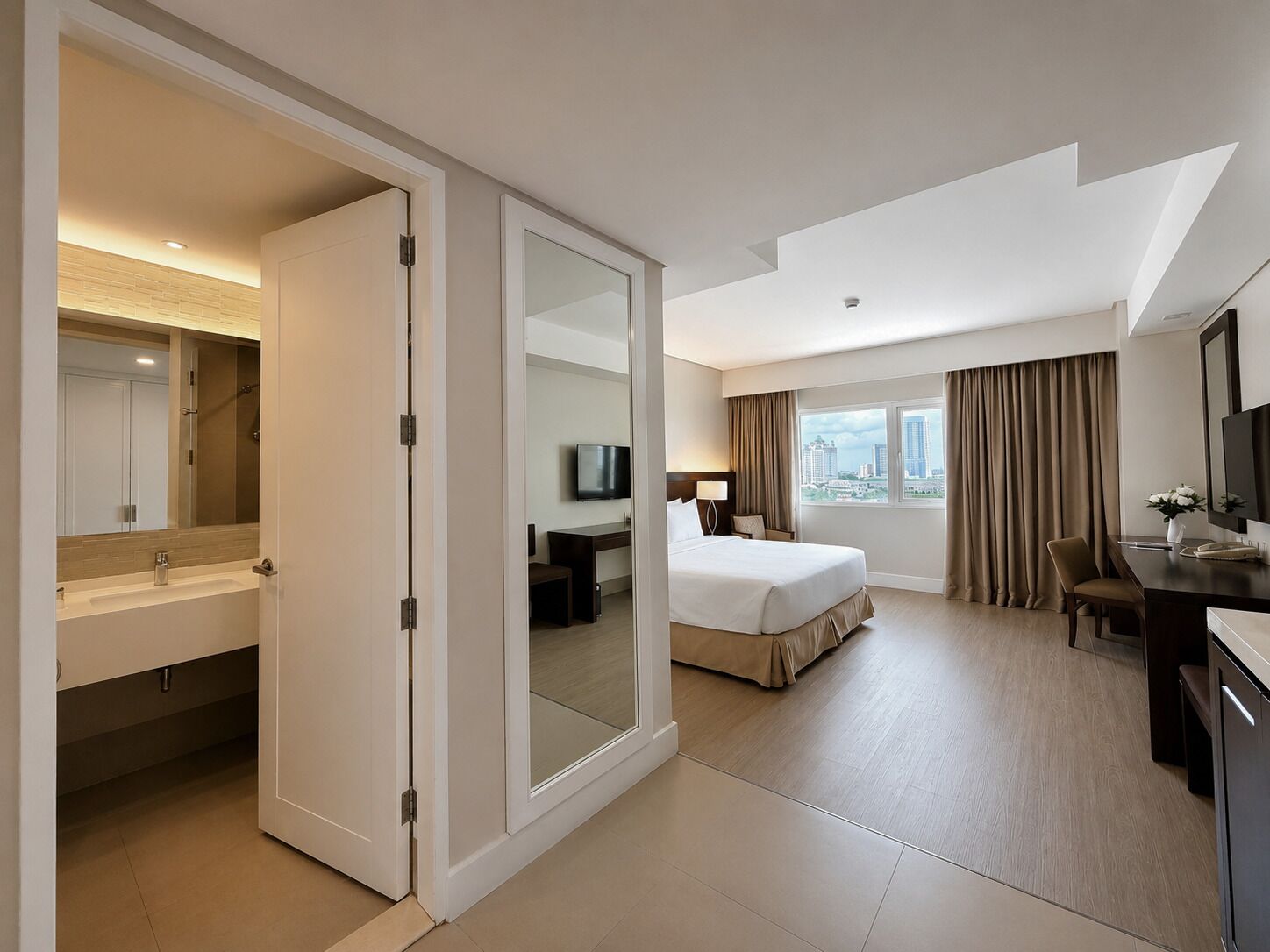 A hotel room with a bed, a desk, a TV, a bathroom with a mirror, and a city view.