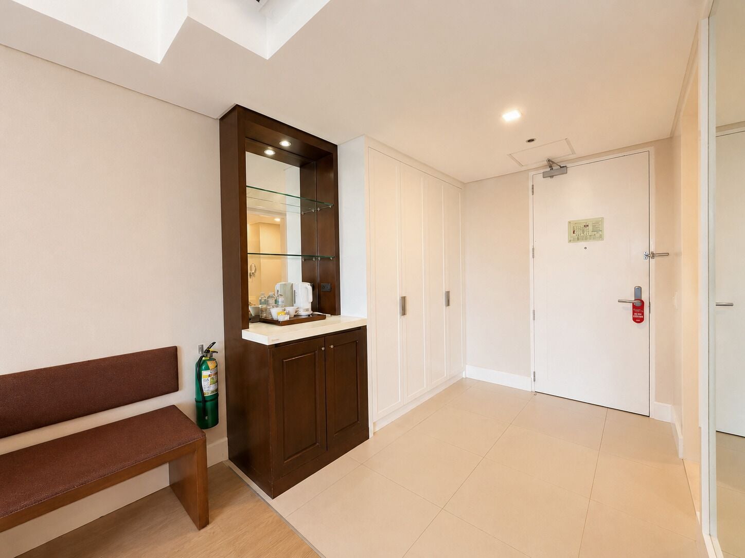 A hotel corridor with a bench, a glass-fronted cabinet with a coffee maker, and a door with a fire alarm.