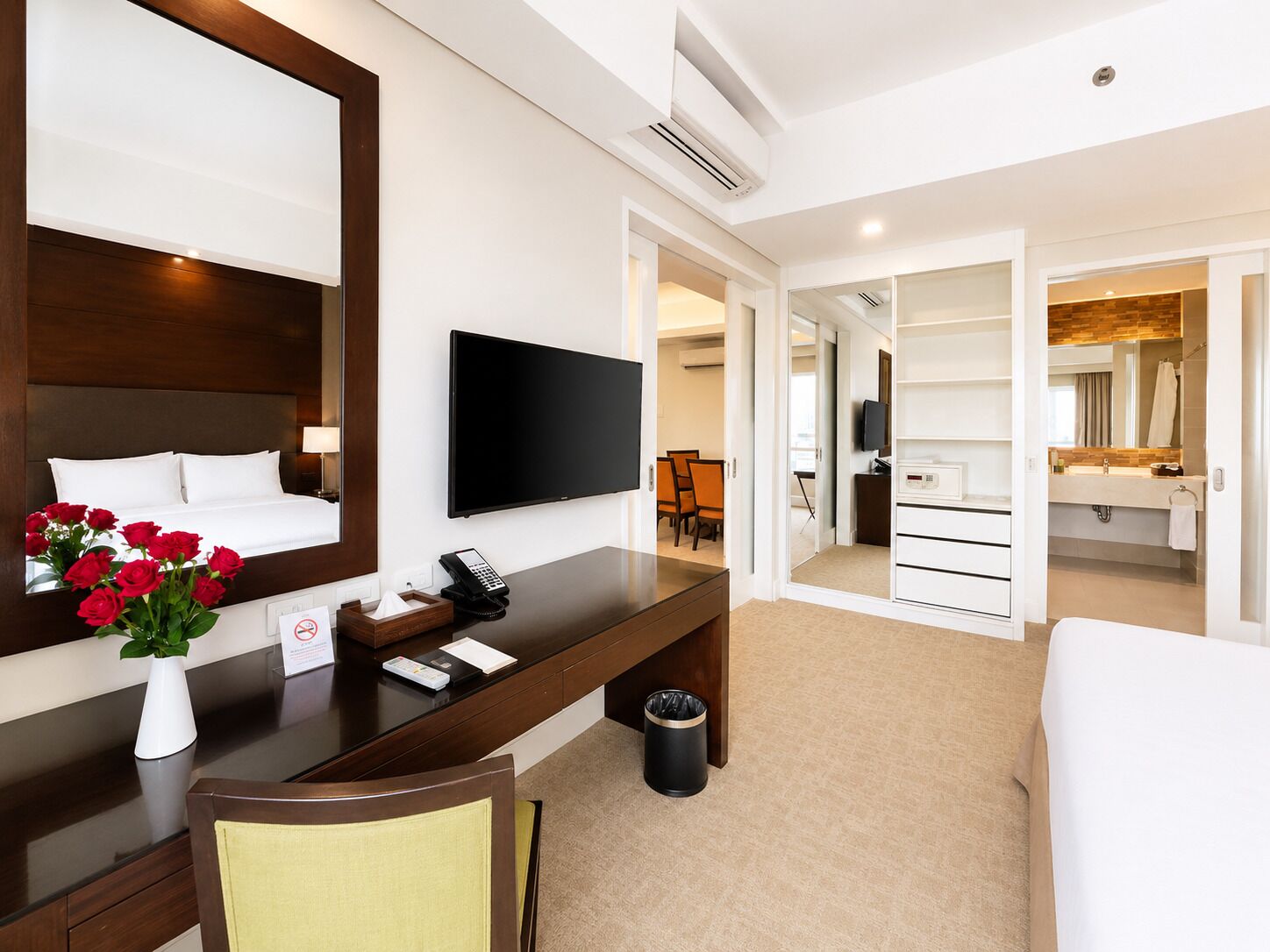 A hotel room with a bed, a desk with a telephone, a flat-screen TV, a mirror, a vase with flowers, and a trash bin.