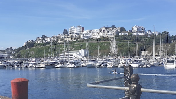 A marina with numerous boats docked, a Ferris wheel, and buildings on a hillside in the background. - The Briarfields (Torquay)