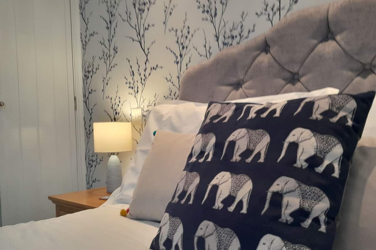 Un dormitorio con cabecera capitonada, almohada estampada, lámpara de noche y pared cubierta de papel tapiz floral.