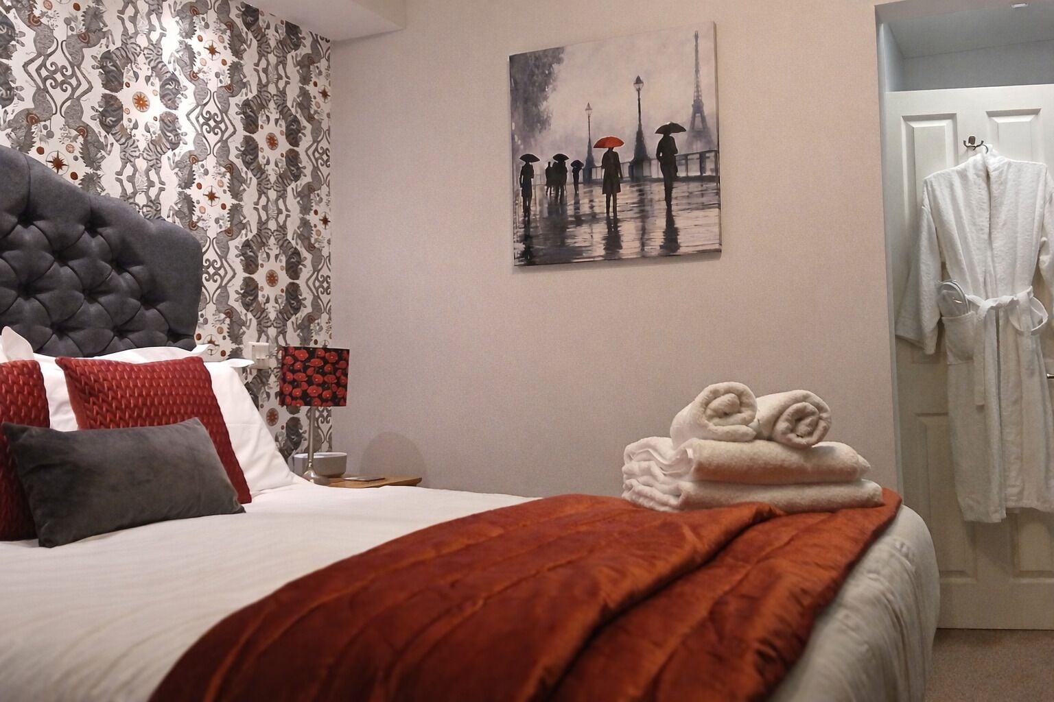 Una habitación de hotel con una cama, un cuadro de personas bajo la lluvia y una lámpara, además de un perchero para abrigos.