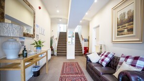 A spacious living area with a leather sofa, a wooden coffee table, and a red patterned rug. A staircase leads to an upper floor, and a framed landscape painting adorns the wall. - The Briarfields (Torquay)