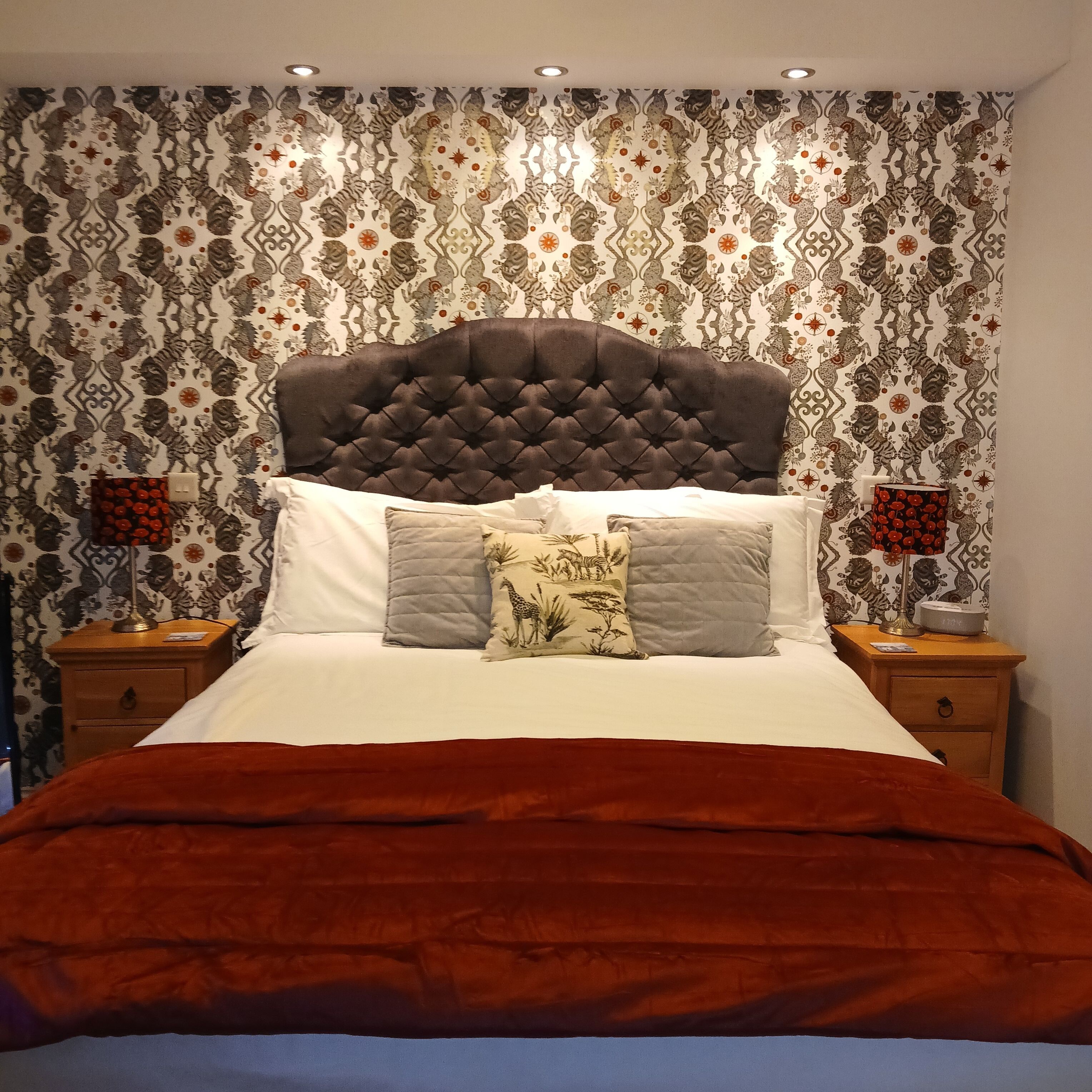 A bed with a tufted headboard, multiple pillows, and a red blanket. There are two bedside tables with lamps and a decorative wall with a patterned wallpaper.