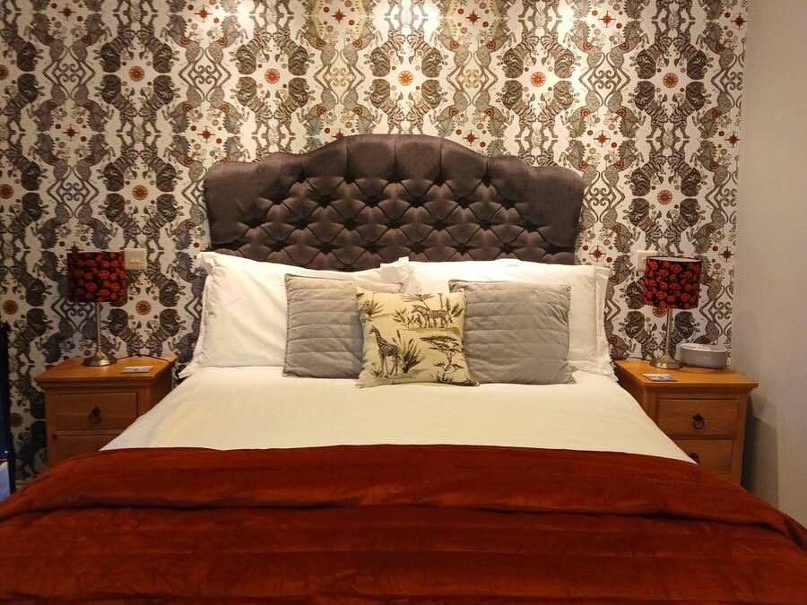 A bed with a tufted headboard, multiple pillows, and a red blanket. There are two bedside tables with lamps and a decorative wall with a patterned wallpaper.