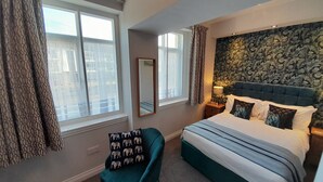 A bedroom with a large bed, a chair, a mirror, and a window with curtains. - The Briarfields (Torquay)